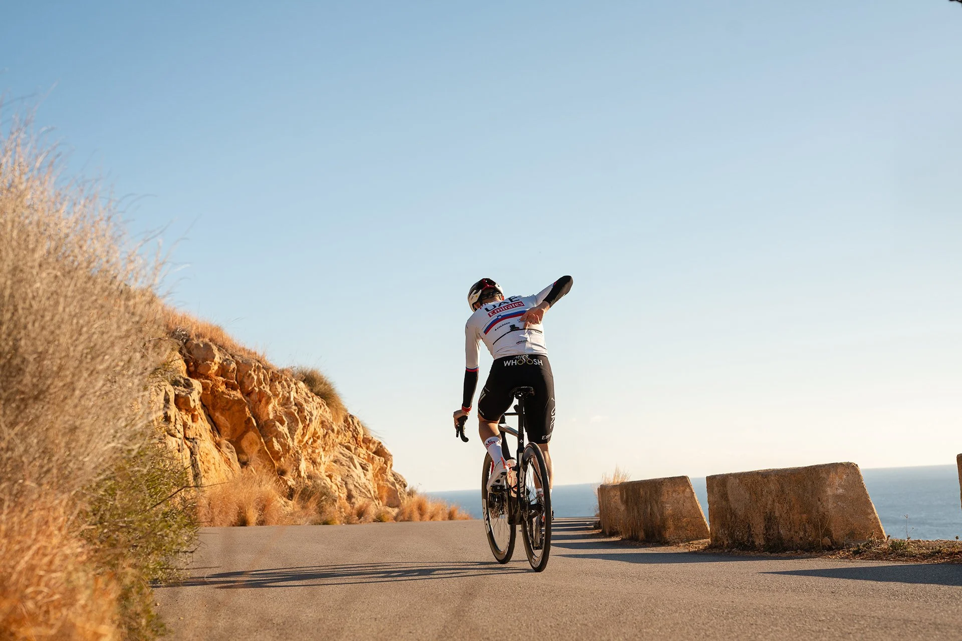 Un ciclista pedala lungo una strada costiera soleggiata con rocce e vegetazione secca a sinistra e il mare visibile in lontananza.