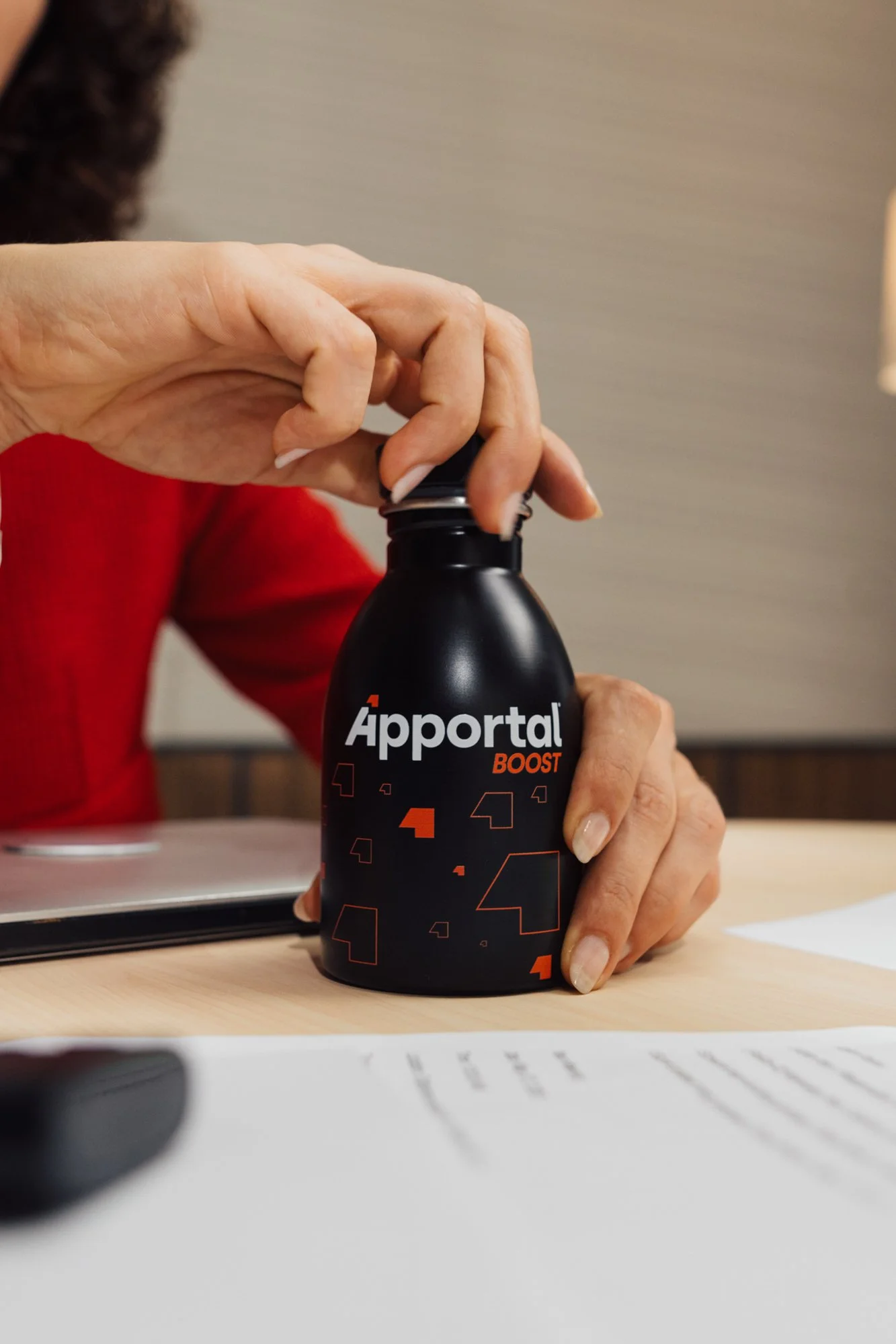 Una persona apre una bottiglia nera con la scritta 'Àportál BOOST' sopra un tavolo di legno, con documenti e un telefono vicino.