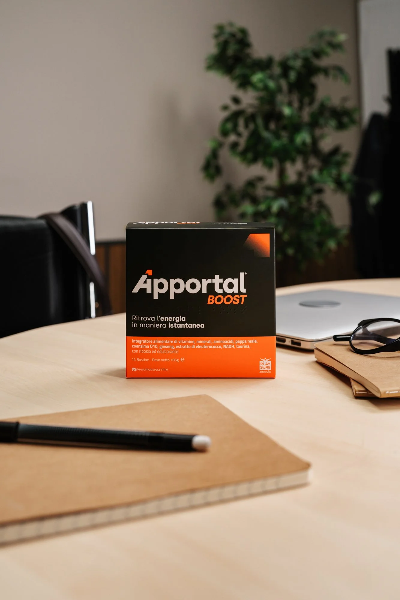 Scatola di integratori chiamata 'Ápportal BOOST' posta su un tavolo di legno, con uno sfondo di parete beige, un albero e alcuni oggetti come un laptop, un notebook, occhiali e una penna sui posti intorno.