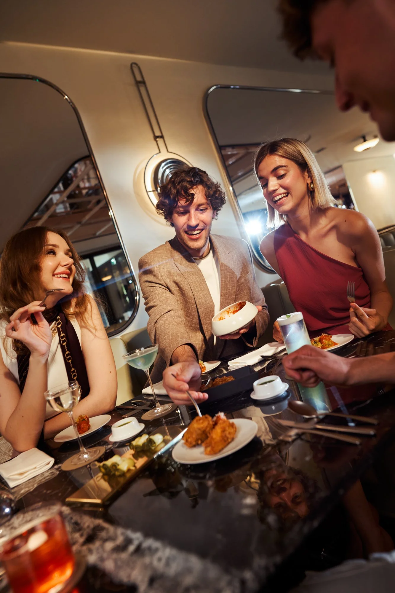 Gruppo di amici che mangiano e brindano in un ristorante elegante, con cibo e cocktail sul tavolo.