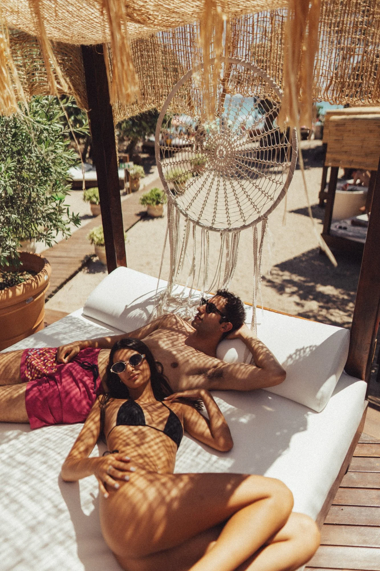 Due persone, una donna in bikini nero e rosa e un uomo in costume da bagno, riposano su una lettino da sole sotto un ombrellone di paglia in un ambiente estivo soleggiato con piante e altre persone sullo sfondo.