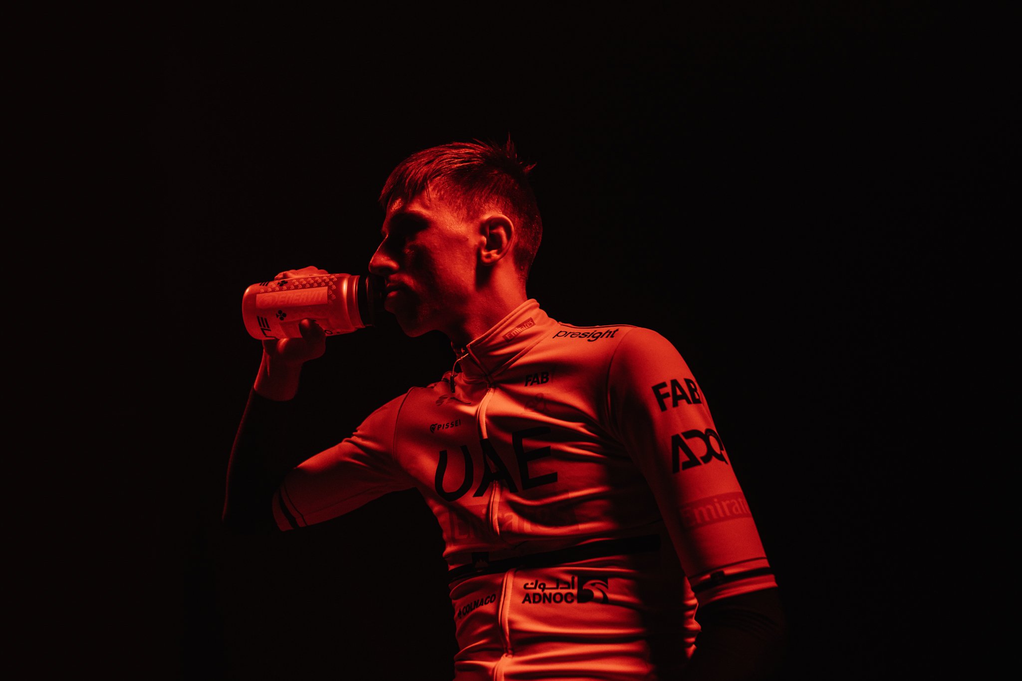 Un ciclista professionista che beve da una borraccia, indossa un completo sportivo e si trova in un ambiente scuro con luci rosse