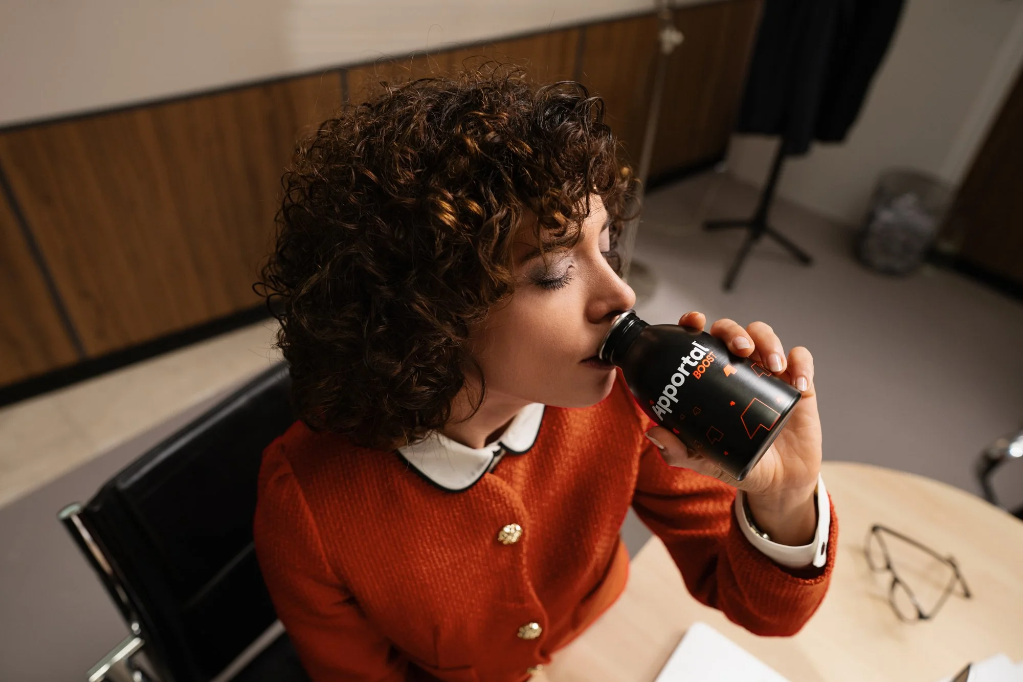 Una donna con capelli ricci bruni beve da una lattina di bevanda energetica in un ambiente interno.