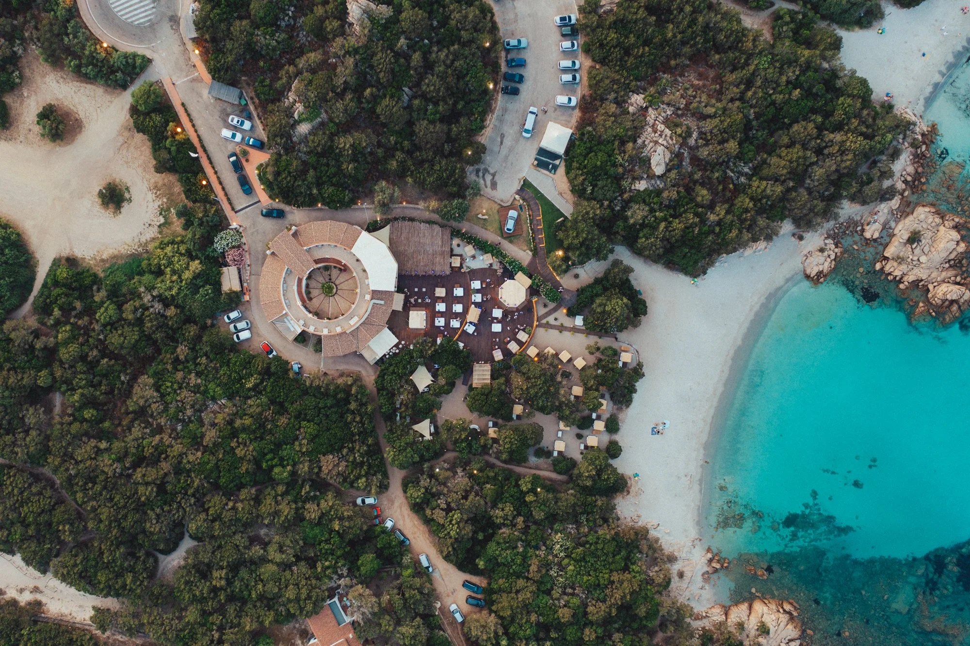 Veduta aerea di una spiaggia con acque turchesi, un'area alberata e un complesso di strutture circolari vicino alla spiaggia, con parcheggi a fianco e sentieri che attraversano la vegetazione.