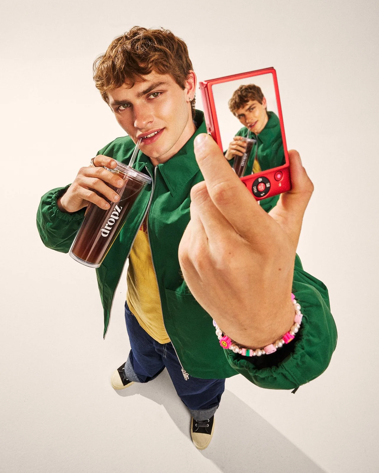 Persona giovane con capelli ricci, indossa giacca verde, pantaloni larghi e scarpe scure, mentre scatta una selfie con una fotocamera rossa e tiene un bicchiere di cola.