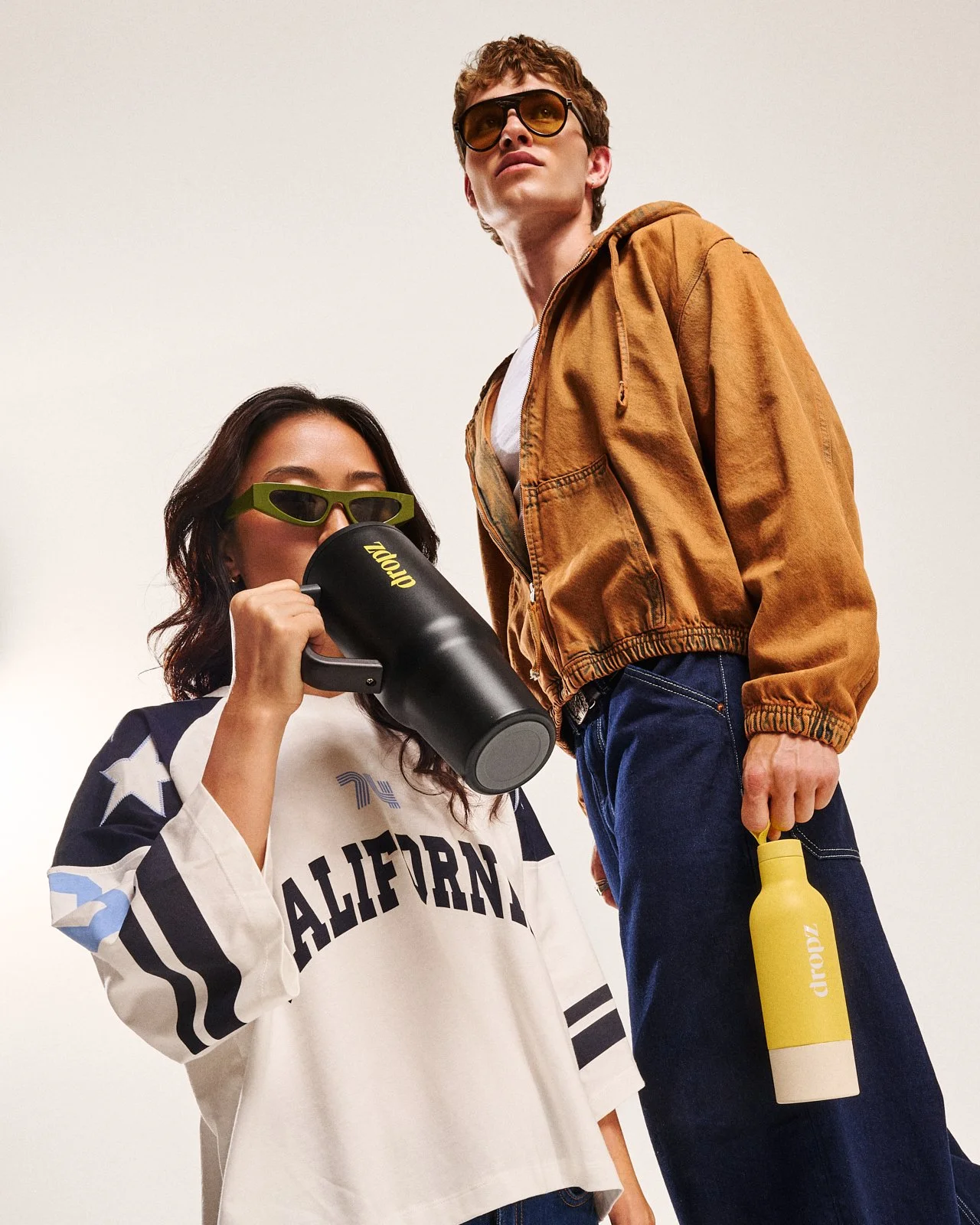 Due giovani posano, uno con una maglietta con scritto "California" che beve da una borraccia nera, l'altra con una giacca marrone e occhiali da sole. Entrambi indossano occhiali fashion e sfoggiano uno stile casual.