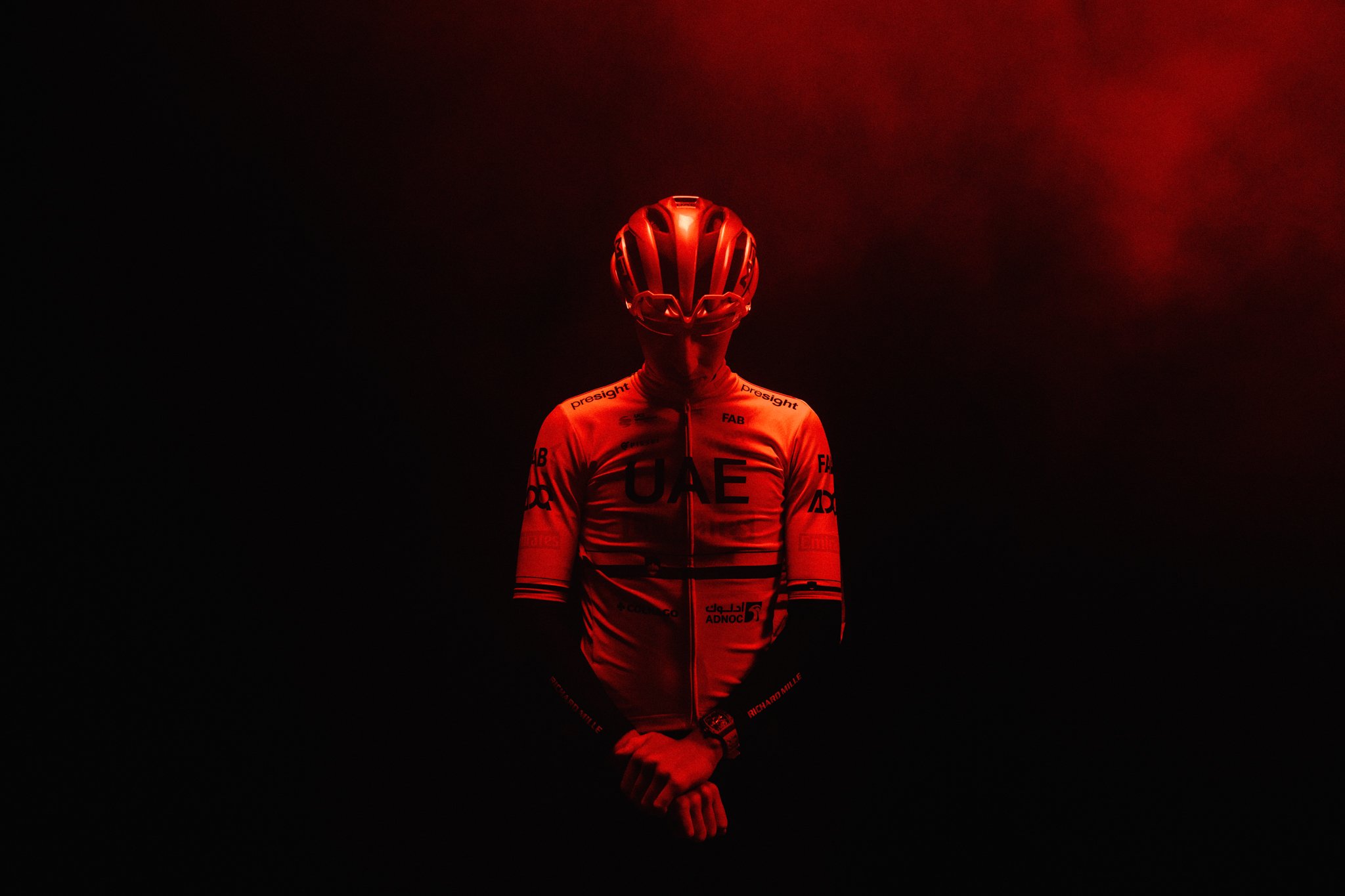 Un ciclista con casco e maglia rossa si staglia contro uno sfondo scuro con luci rosse, creando un effetto visivo drammatico.