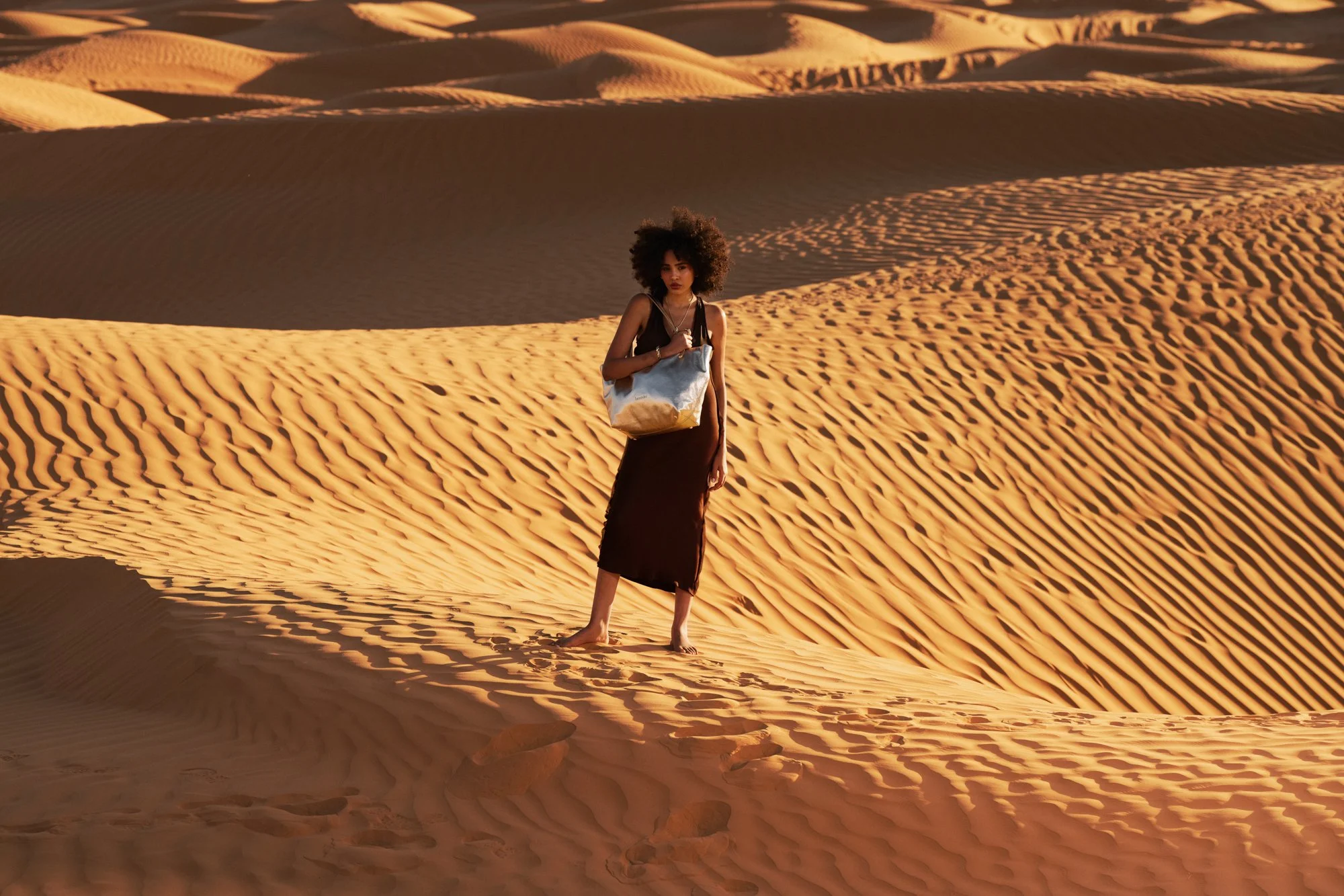 Una donna con capelli afro e vestito nero cammina in un deserto di dune di sabbia con un sacco sulle spalle, rondini volano nel cielo.
