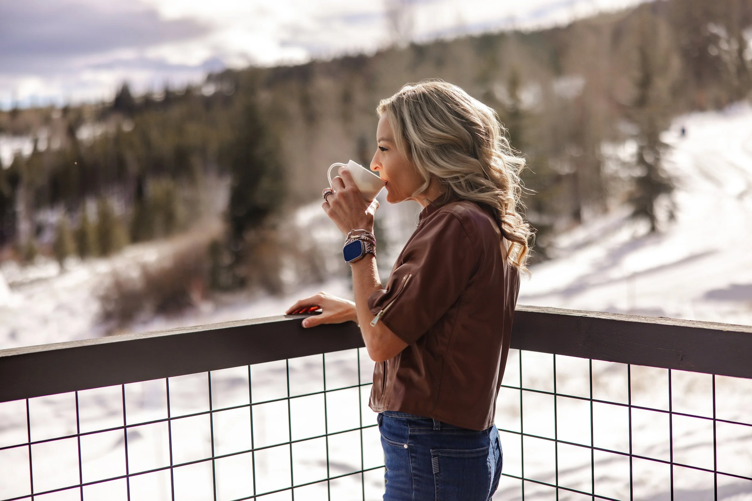 A woman with blonde curly hair drinking from a mug outdoors in a snowy landscape, wearing a brown jacket and blue jeans, standing on a balcony with a metal railing.