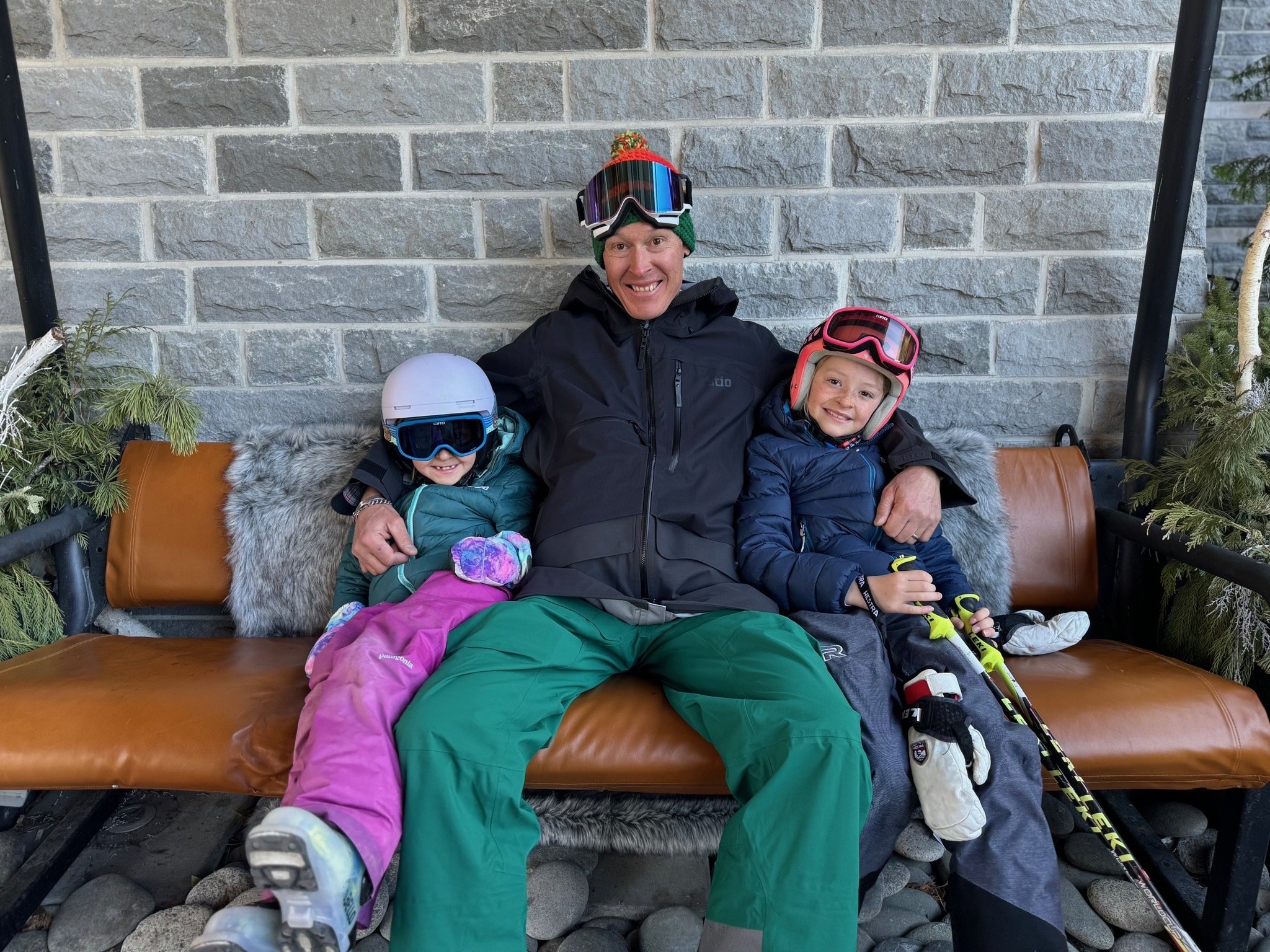A man and two children sitting on a bench outdoors, all wearing ski helmets and winter jackets, smiling at the camera. The man is in the middle, with the children on either side.