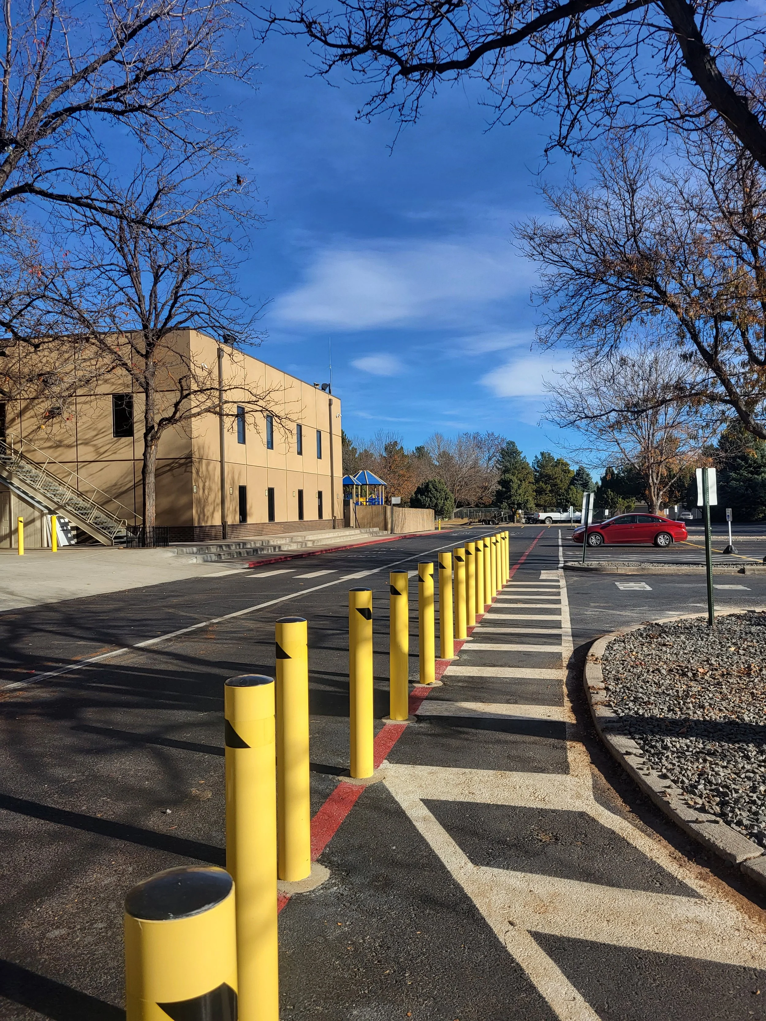 Empty parking lot with yellow safety posts, a red car, leafless trees, a beige building, and a bright blue sky with a few clouds.