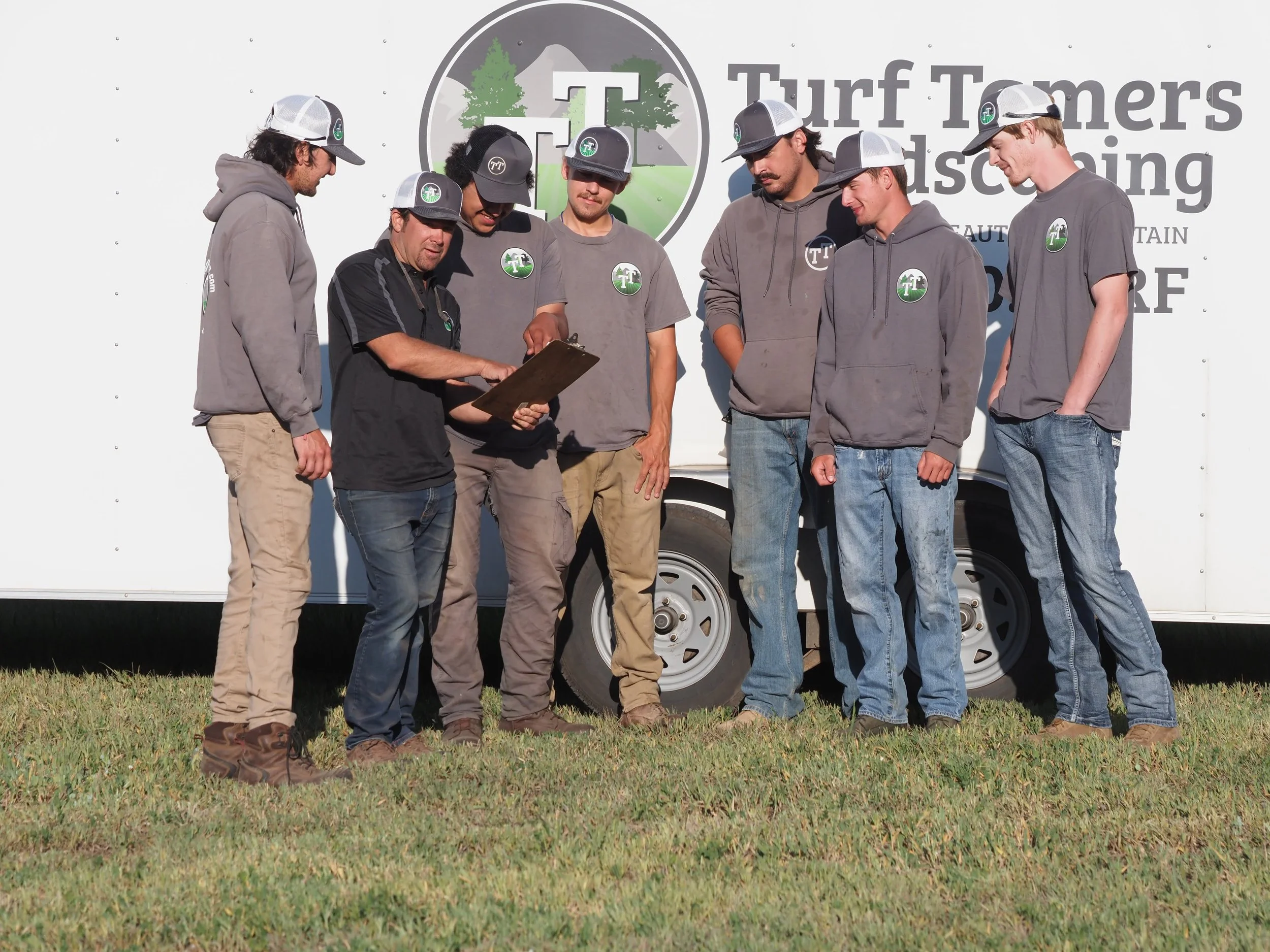 Group of young men and an older man standing outdoors, looking at a clipboard in front of a white truck with a sign that says Turf Tamers Landscaping.