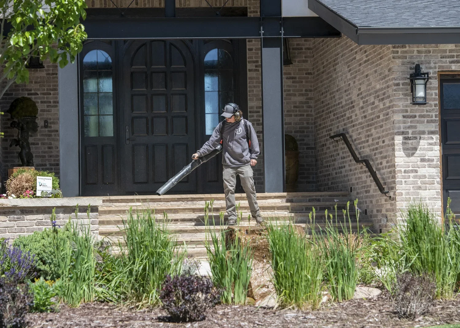 A person wearing a gray hoodie, khaki pants, and a baseball cap is using a leaf blower to clear outside stairs in front of a brick house with a black door and steps, surrounded by greenery and landscaping.