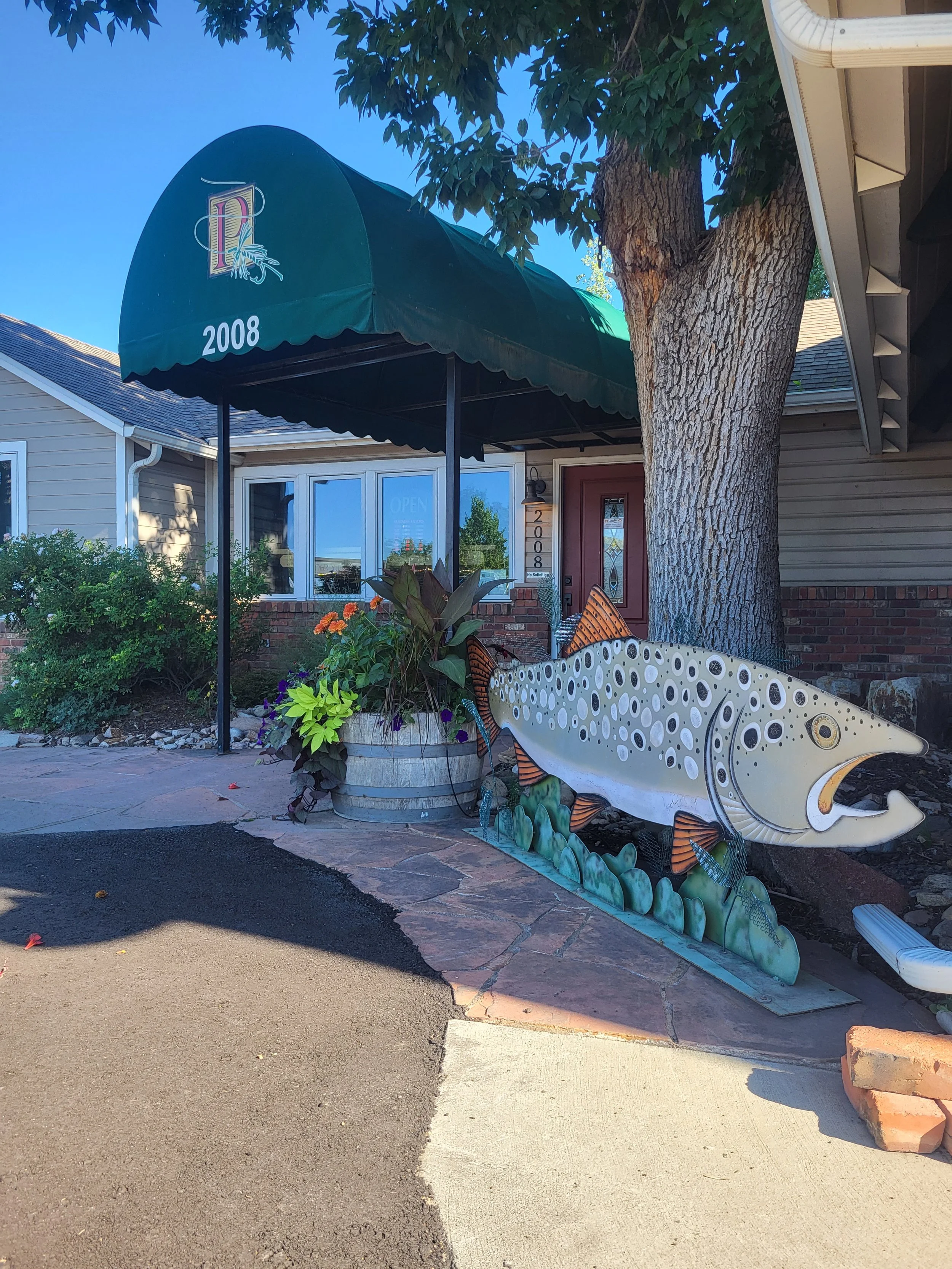 Decorative outdoor sculpture of a fish placed in front of a house with a large tree, potted plants, and a green awning that says '2008'.