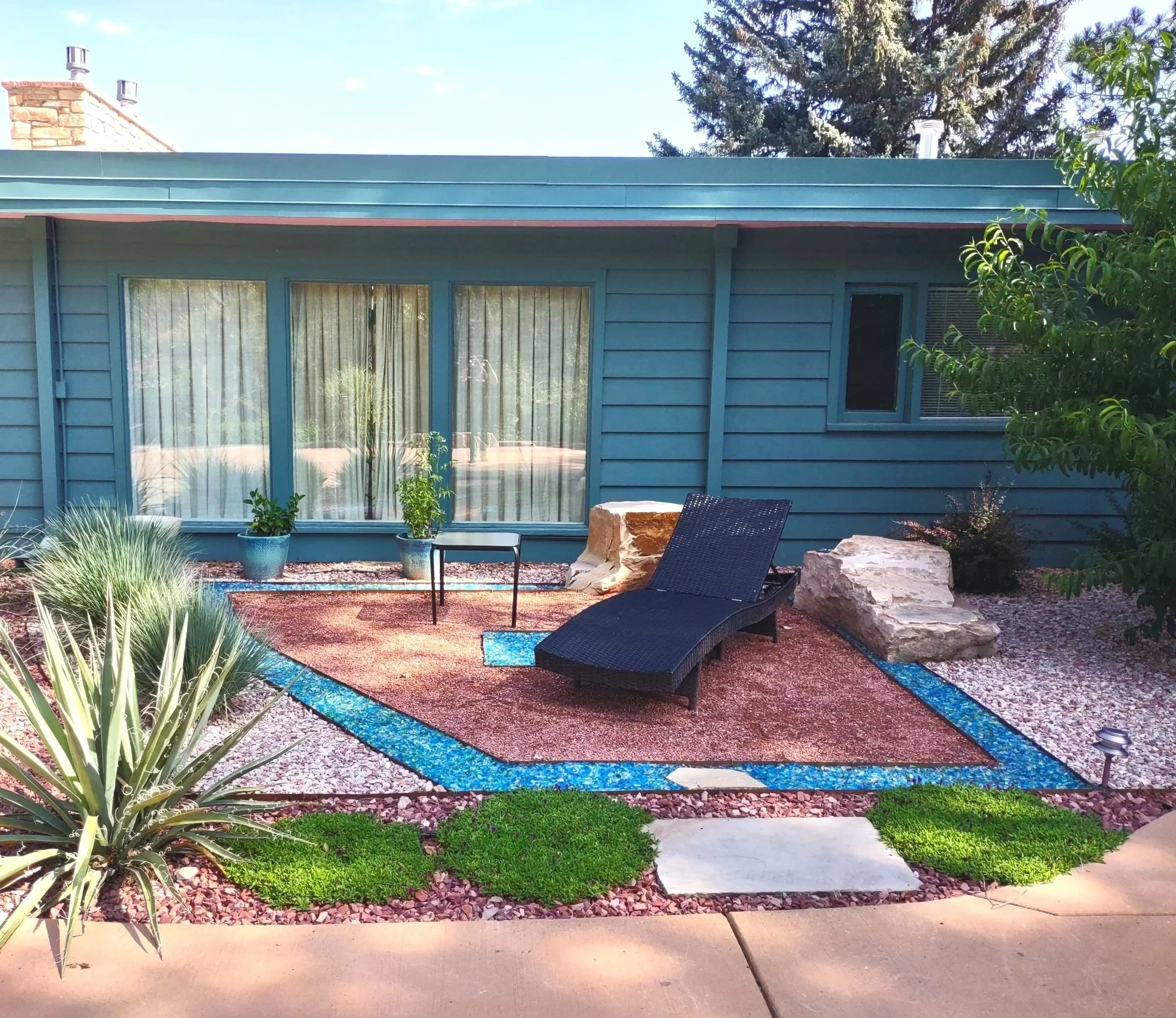Backyard garden scene with patio furniture, colorful pathway, plants, and large window of a house.