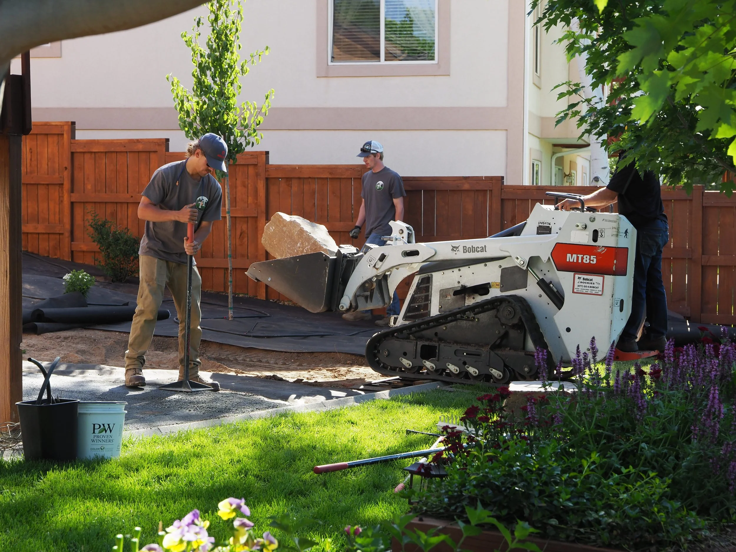 Workers landscaping backyard, using heavy machinery to level ground, surrounded by flowers, trees, and a wooden fence.