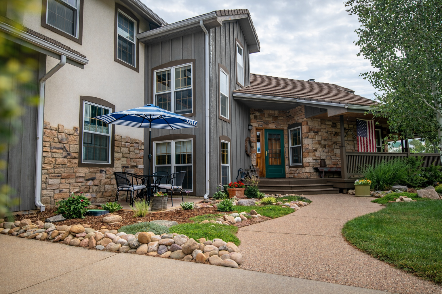 Front yard of a house with a stone and siding facade, a porch with an American flag, and outdoor furniture under a blue and white striped umbrella. The yard has a curved pathway, landscaped with rocks, plants, and flowers.