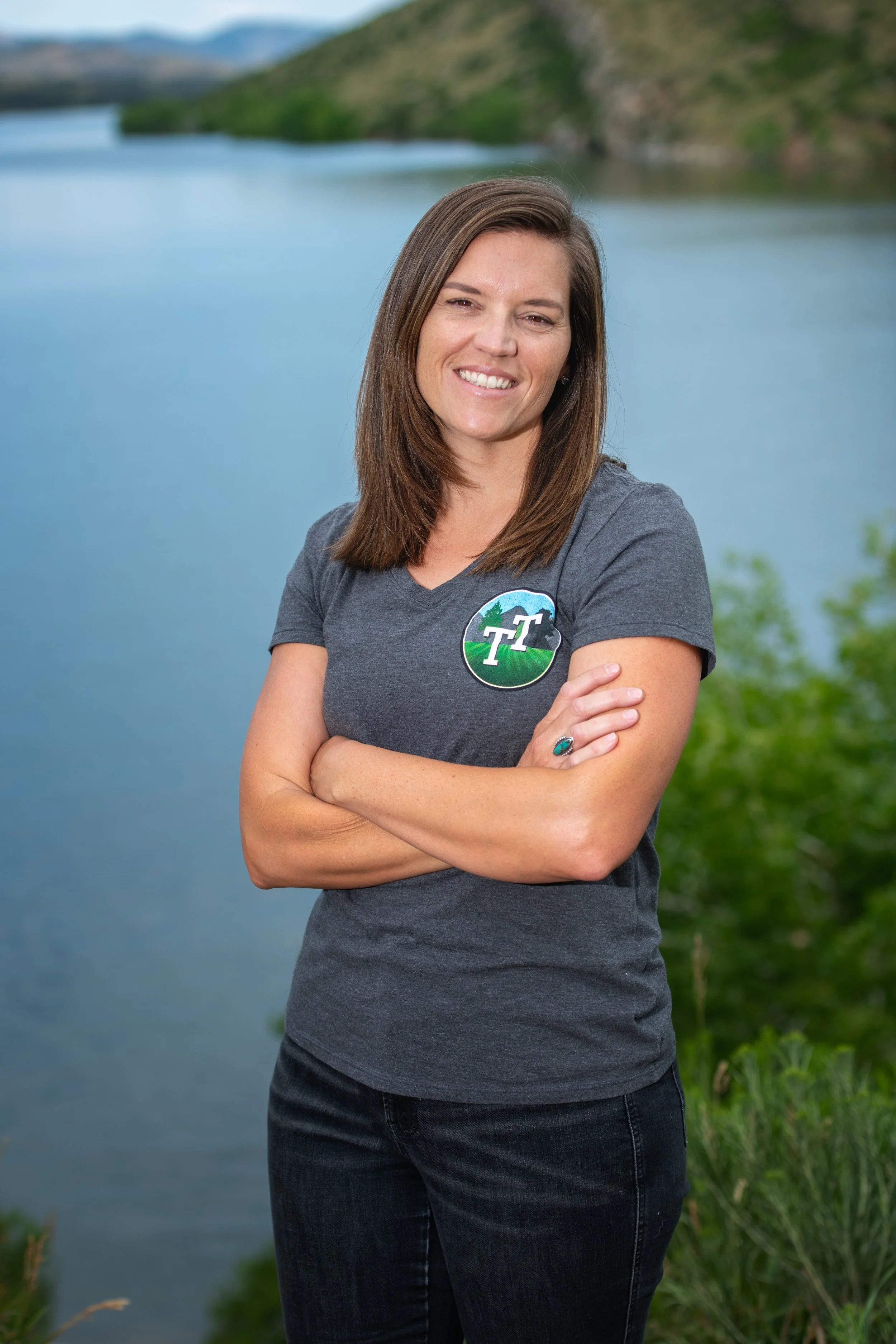 A woman with long brown hair, smiling and crossing her arms, stands outdoors near a body of water with lush green trees and mountains in the background, wearing a gray t-shirt with a logo.