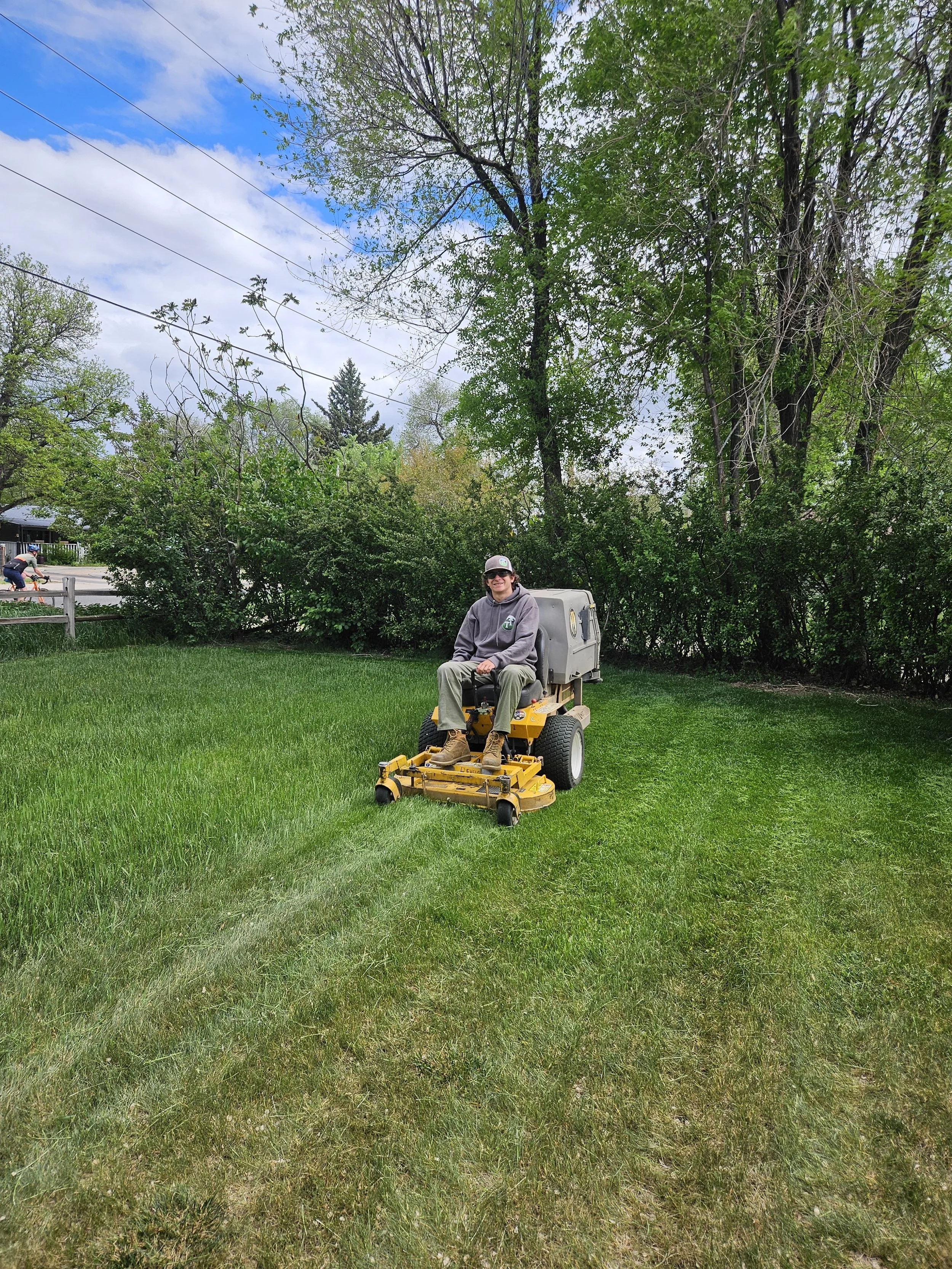 A person riding a yellow riding lawn mower on a green lawn with trees and bushes in the background, under partly cloudy skies.