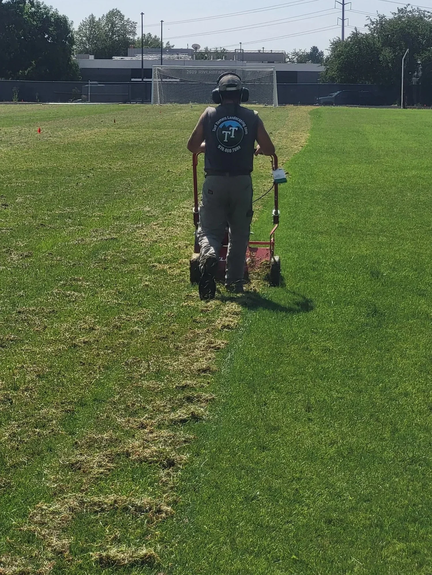 A person wearing a cap, safety ear protection, a sleeveless T-shirt, and pants is mowing or trimming grass on a field with a roller or lawn mower. There is a soccer goal and some buildings and trees in the background.