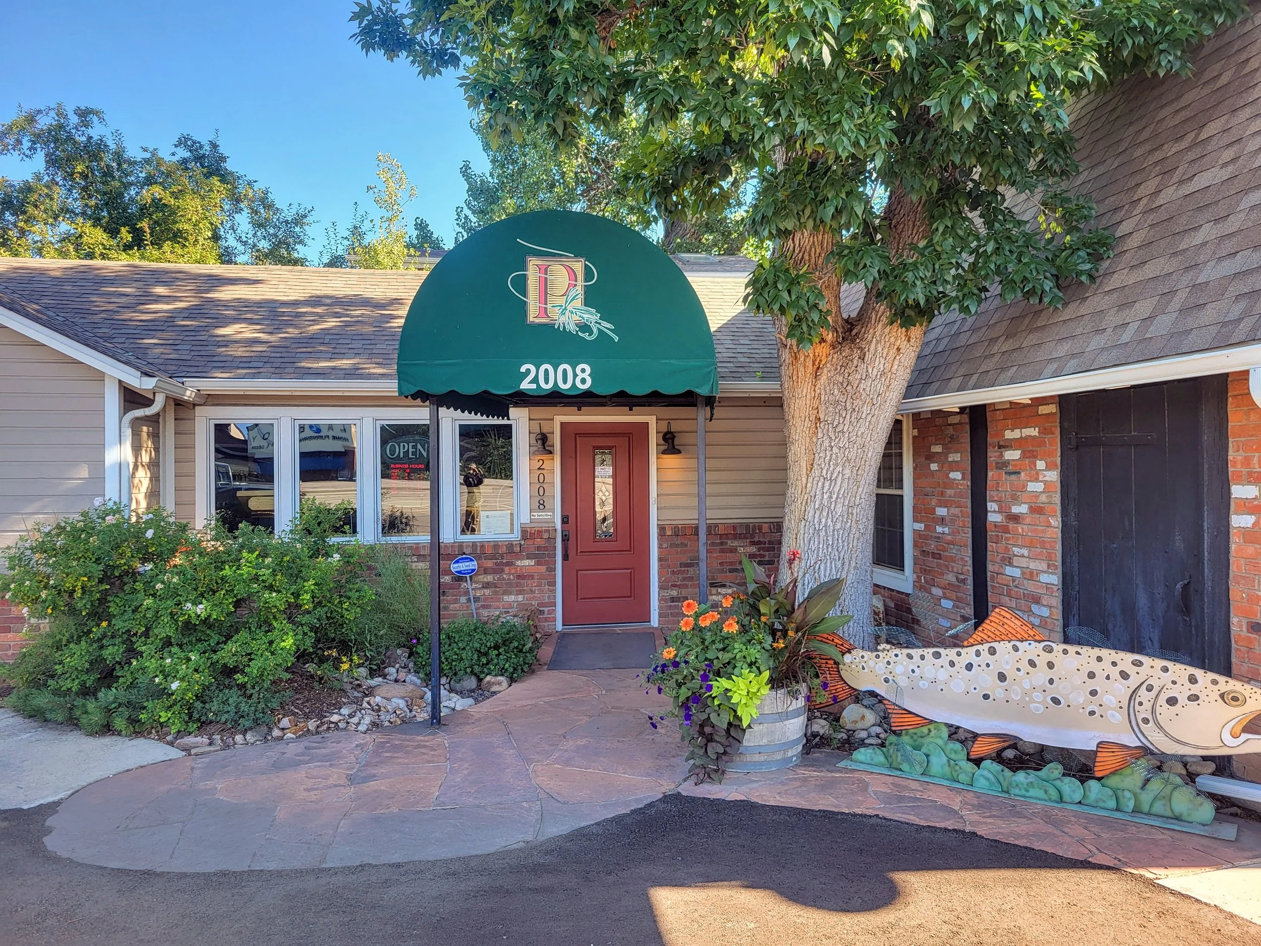 Exterior of a restaurant with a green awning displaying the number 2008 and a logo, a red door, a large tree, colorful plants, and a decorative fish sculpture outside.