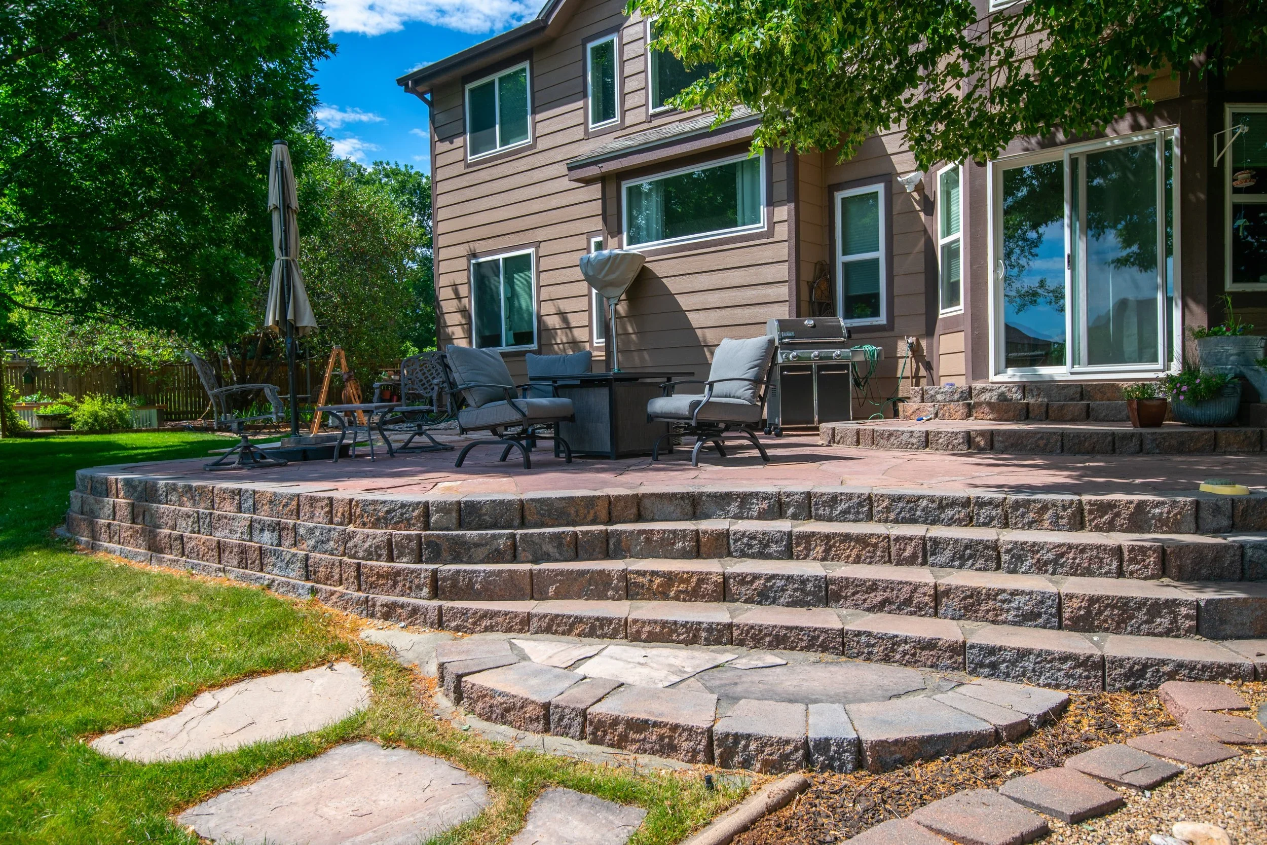 A backyard patio with outdoor furniture, a grill, and potted plants, adjacent to a house with large windows and sliding glass door, surrounded by trees and a lawn.