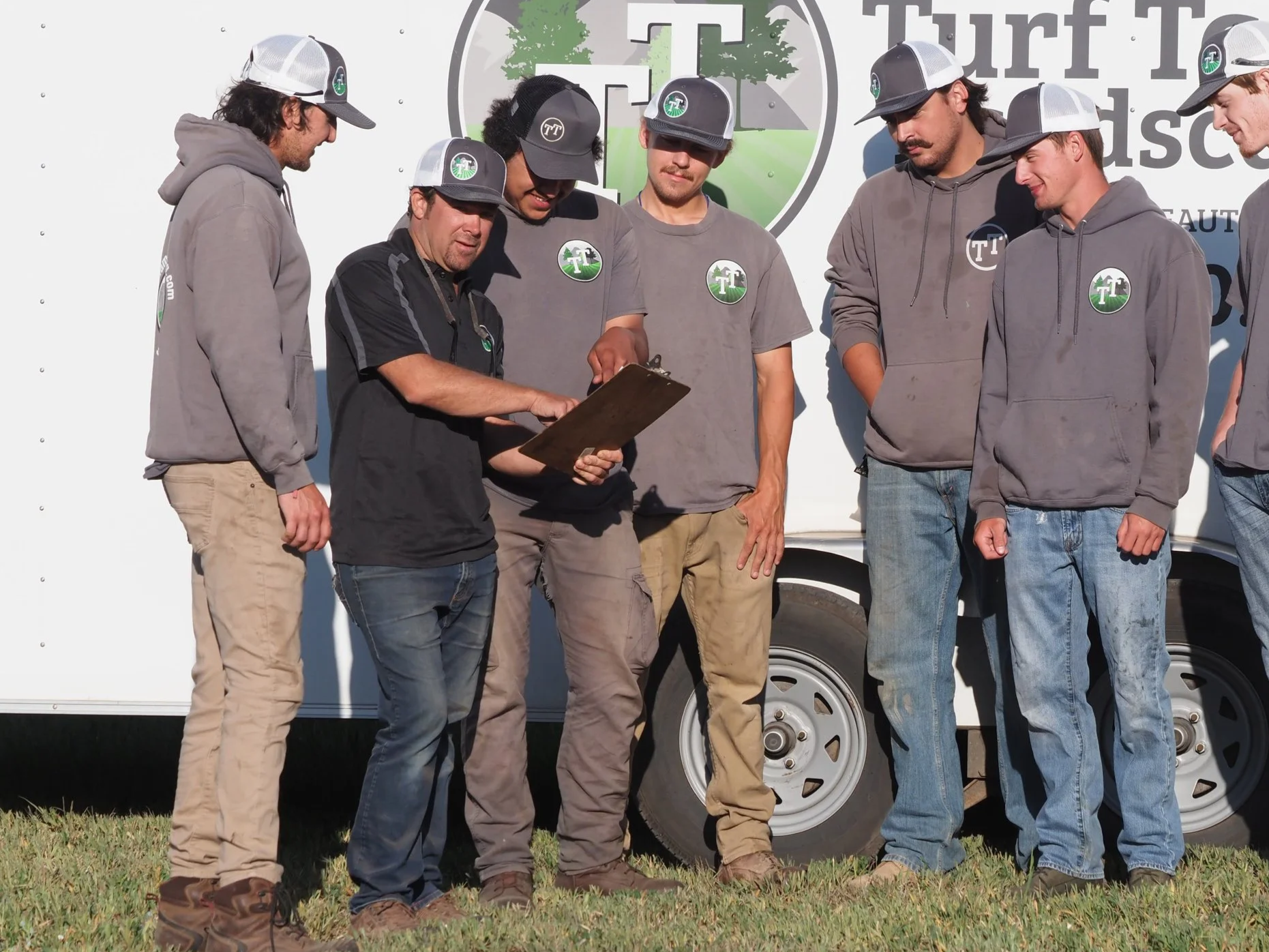 Group of six young men in casual clothing, standing outdoors in front of a white truck with a logo, looking at a clipboard held by one of them. They are wearing gray and black caps, some with logos, and gray hoodies with logos on the chest. The background shows part of a logo and grass on the ground.