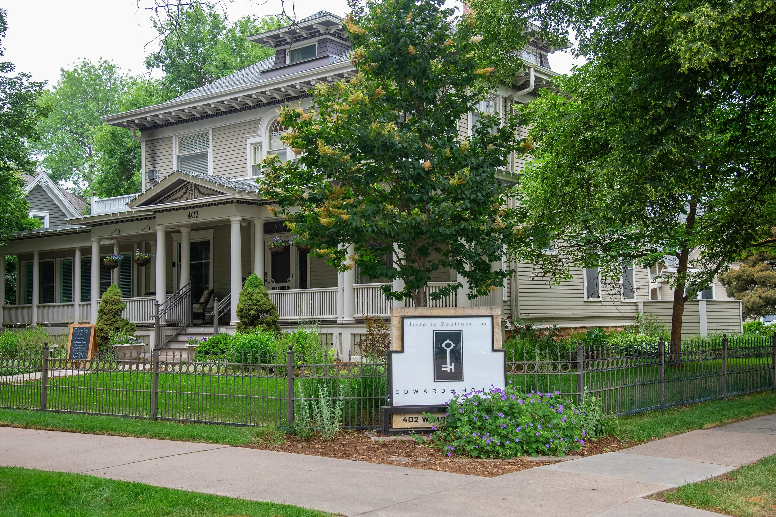 A large historic house with a porch, surrounded by a black fence, green lawn, and trees. A sign reads 'Historic Boutique Inn' with a key and house logo, and the name 'Edward House'.