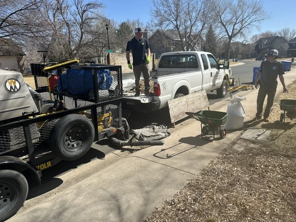 Two men working on a residential street with a white pickup truck, a trailer loaded with equipment, and gardening supplies, including a garden cart and bags of yard waste. The scene appears to be outdoor work or yard cleanup.
