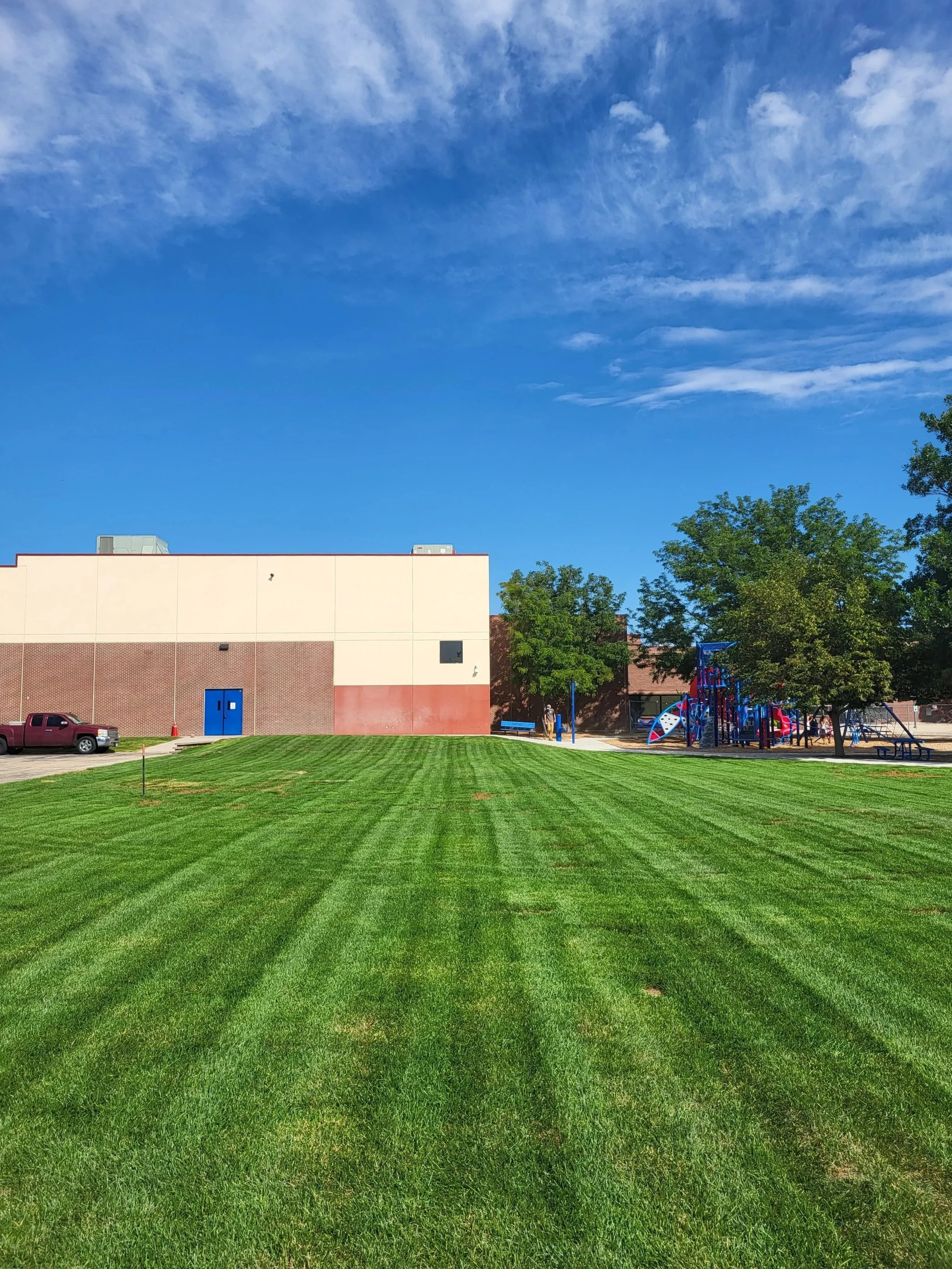 A well-maintained grassy field in front of a school building with a playground on the right side, blue sky with some clouds overhead.