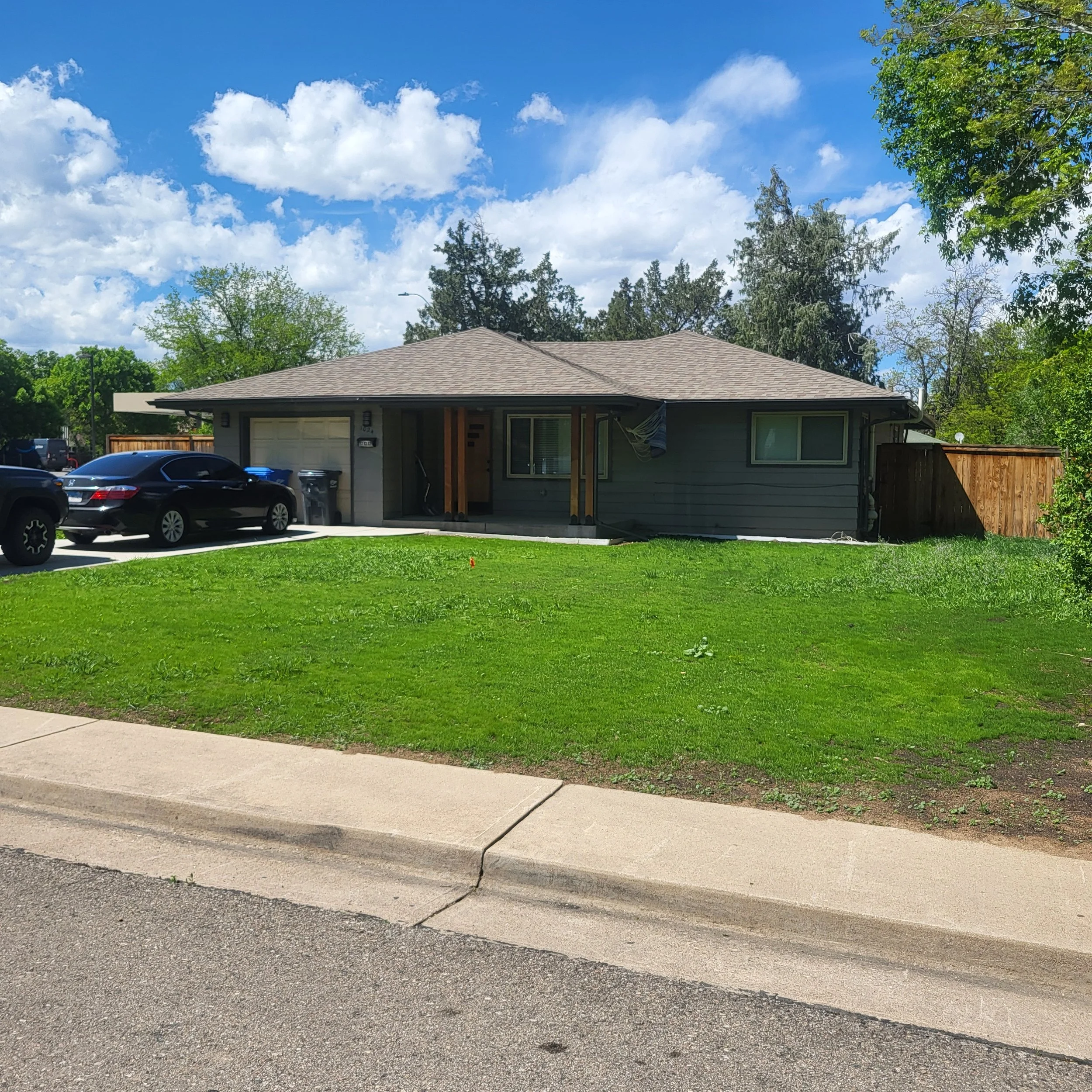 A single-family house with gray siding, a two-car garage, and a front porch. The house has a brown shingled roof, and there are parked cars and trash bins in the driveway. The front yard is grassy, with a sidewalk and street in the foreground, and tr