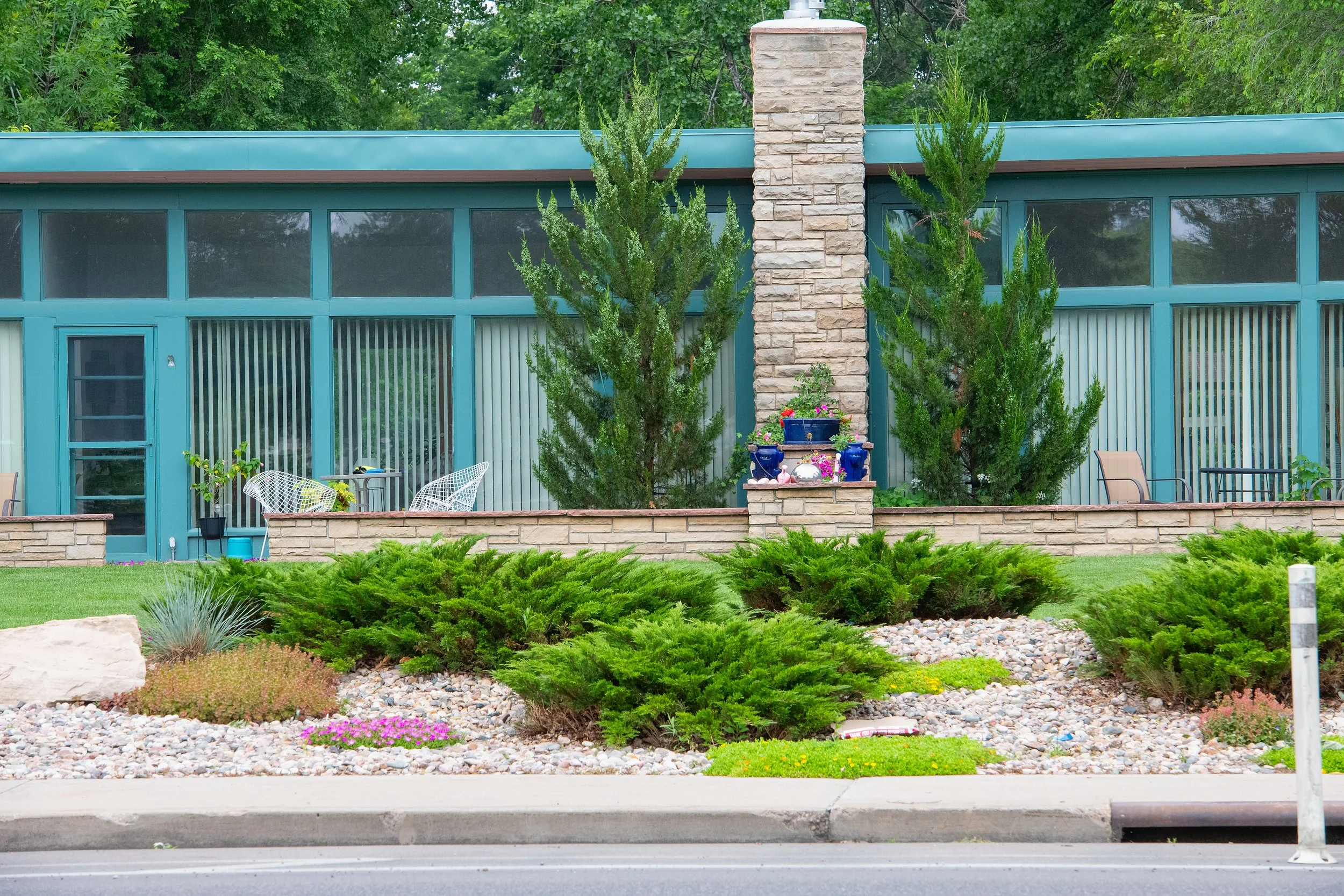 Front view of a mid-century modern house with large glass windows, a brick chimney, and a garden with shrubs, rocks, and trees, with a sidewalk in the foreground.