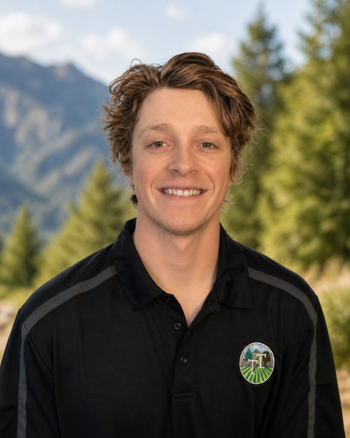 A young man with wavy light brown hair, smiling, wearing a black polo shirt with a patch on the left chest, standing outdoors with a blurred mountain and forest in the background.