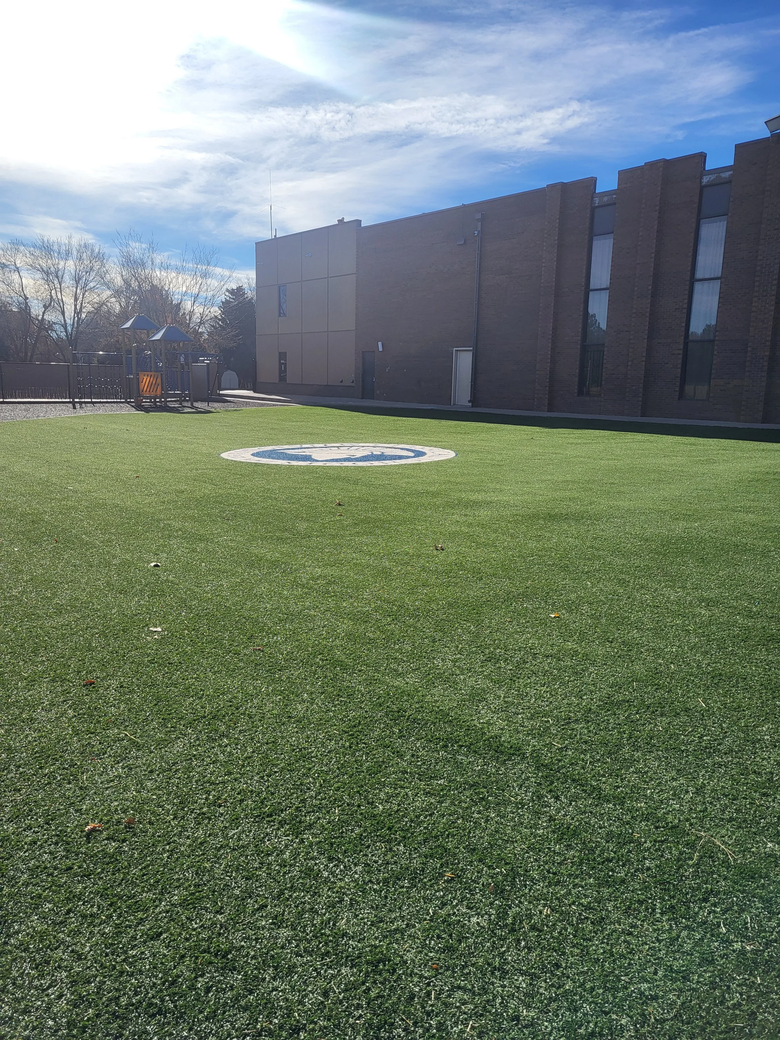 Green artificial turf on a courtyard with a logo in the middle, surrounded by a brick building and a playground in the background under a partly cloudy sky.
