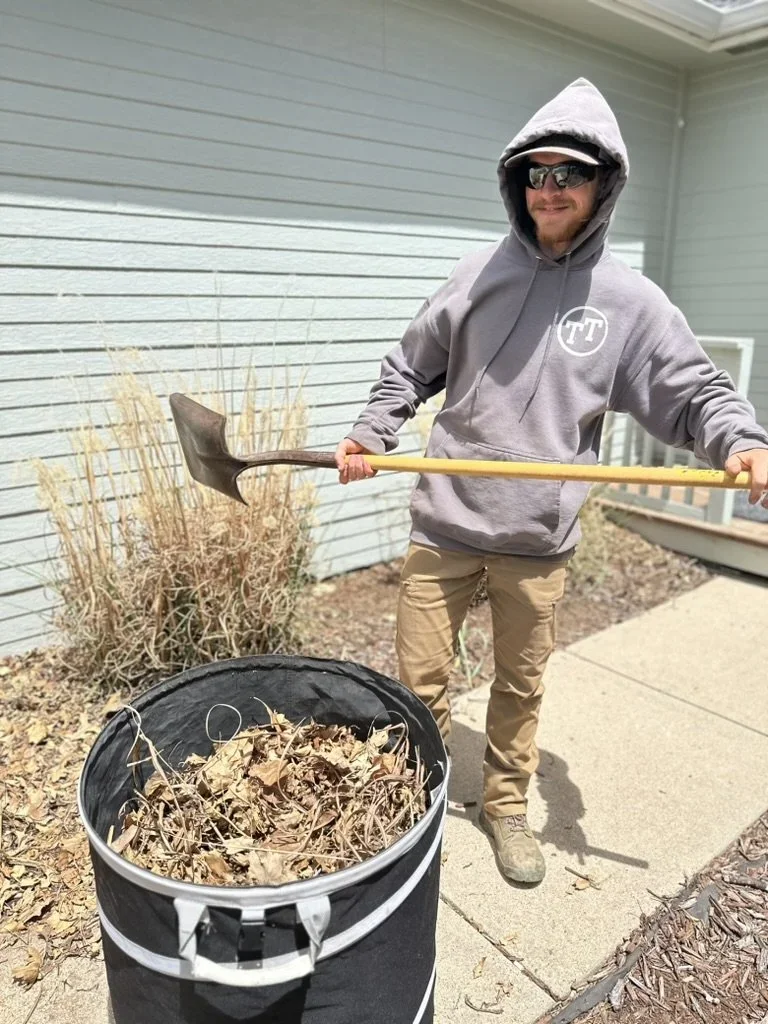 Man in gray hoodie and khaki pants holding a shovel, standing outside near a house, next to a black trash bag filled with dried leaves and garden debris.