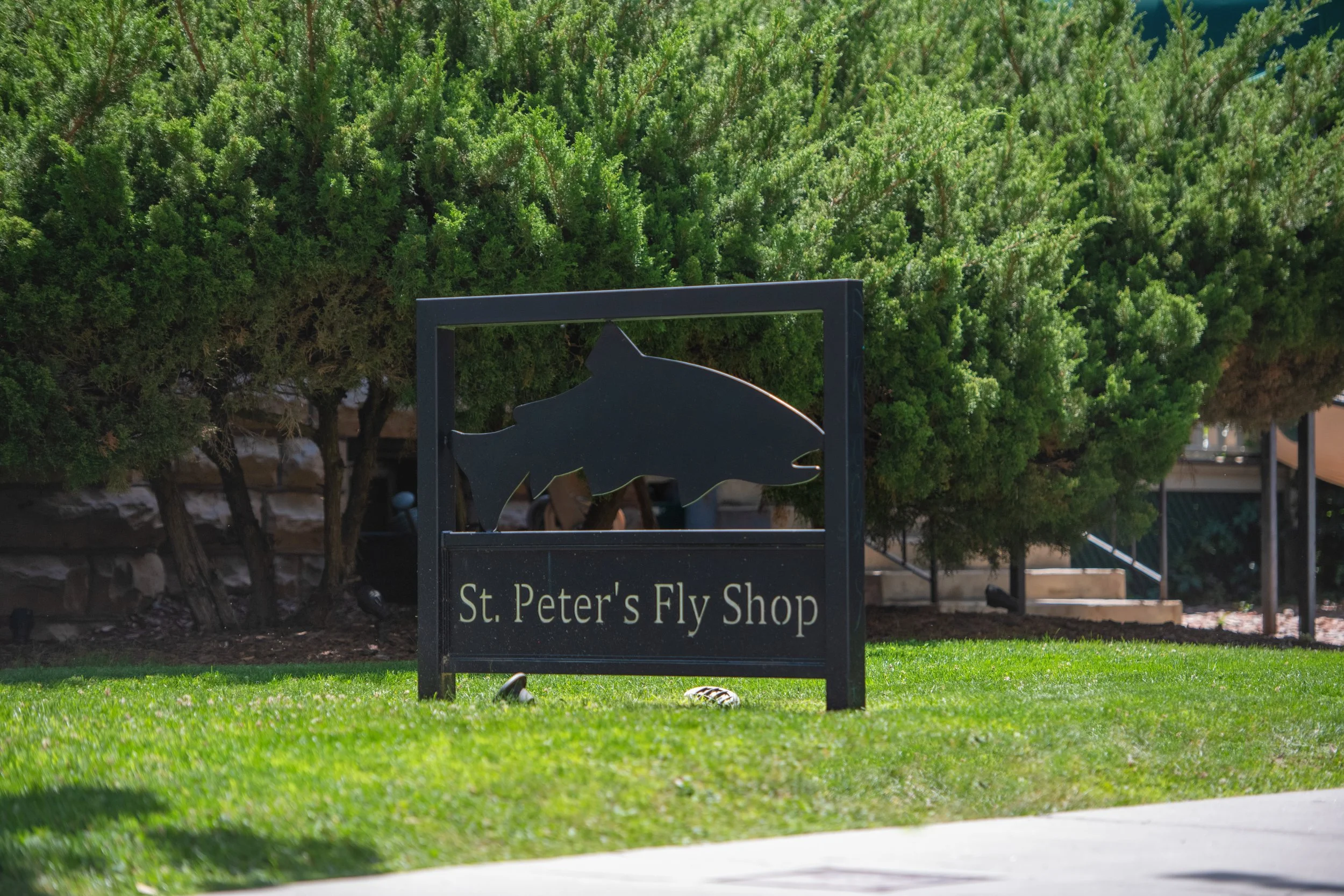 Sign for St. Peter's Fly Shop with a silhouette of a fish, placed on a well-maintained grassy area with bushes and trees in the background.