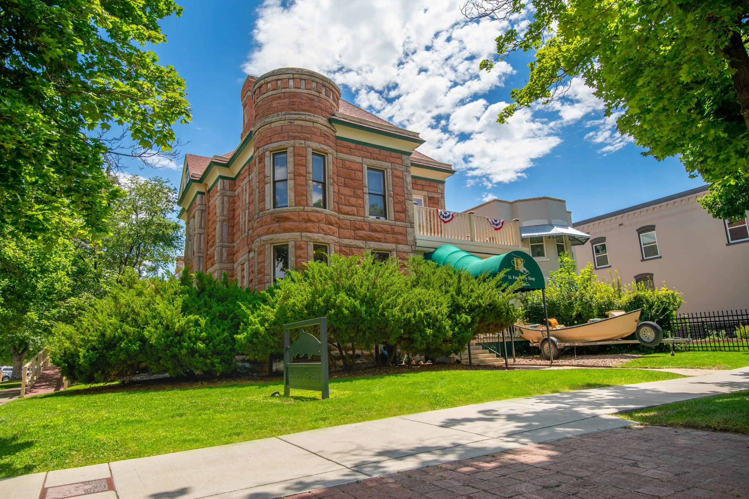 A historic red brick building with arched windows and a turret, surrounded by green trees and bushes, under a partly cloudy blue sky.