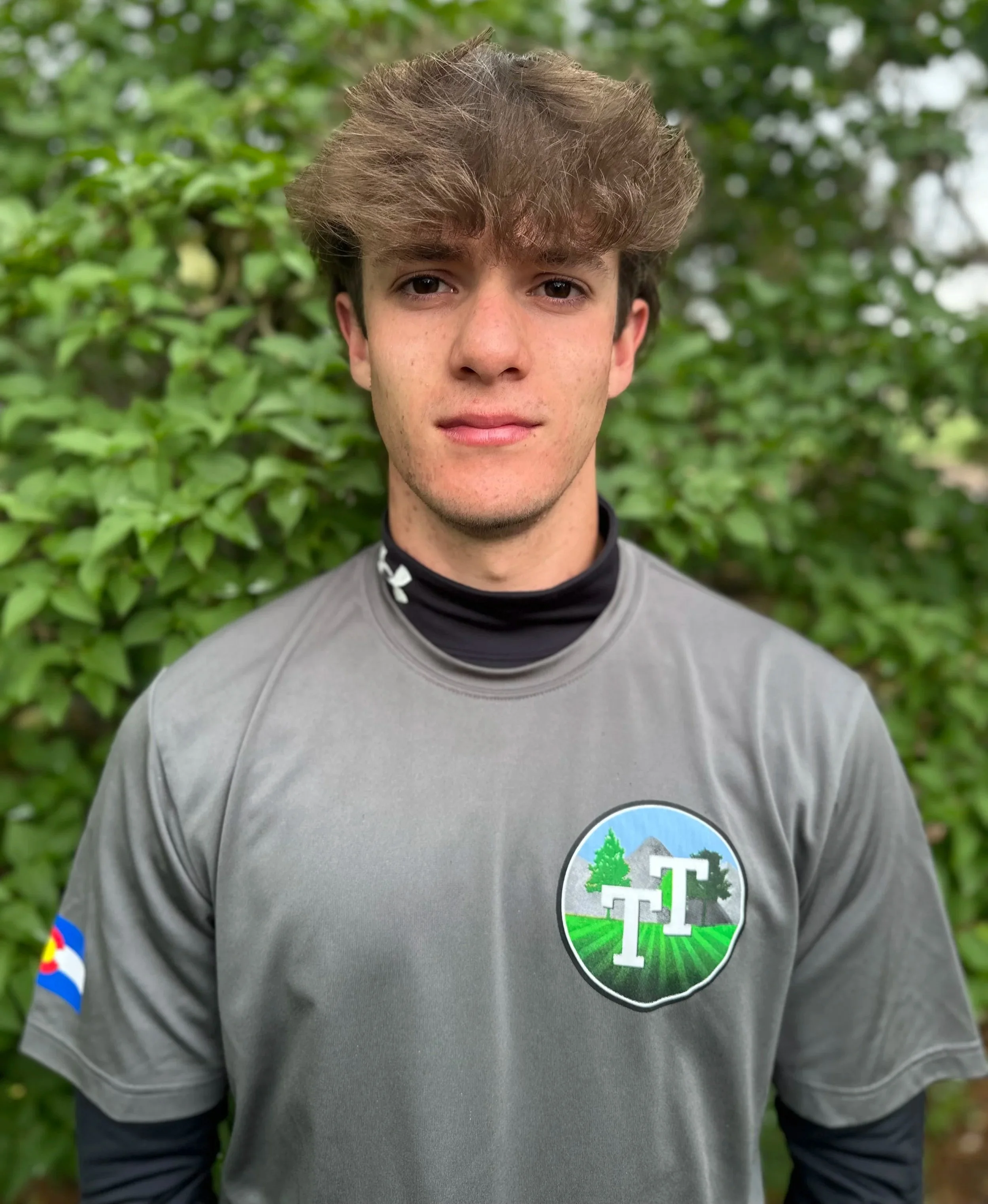 A young man with light skin, tousled brown hair, and freckles wearing a gray shirt and black undershirt, standing outdoors with green leafy bushes in the background.