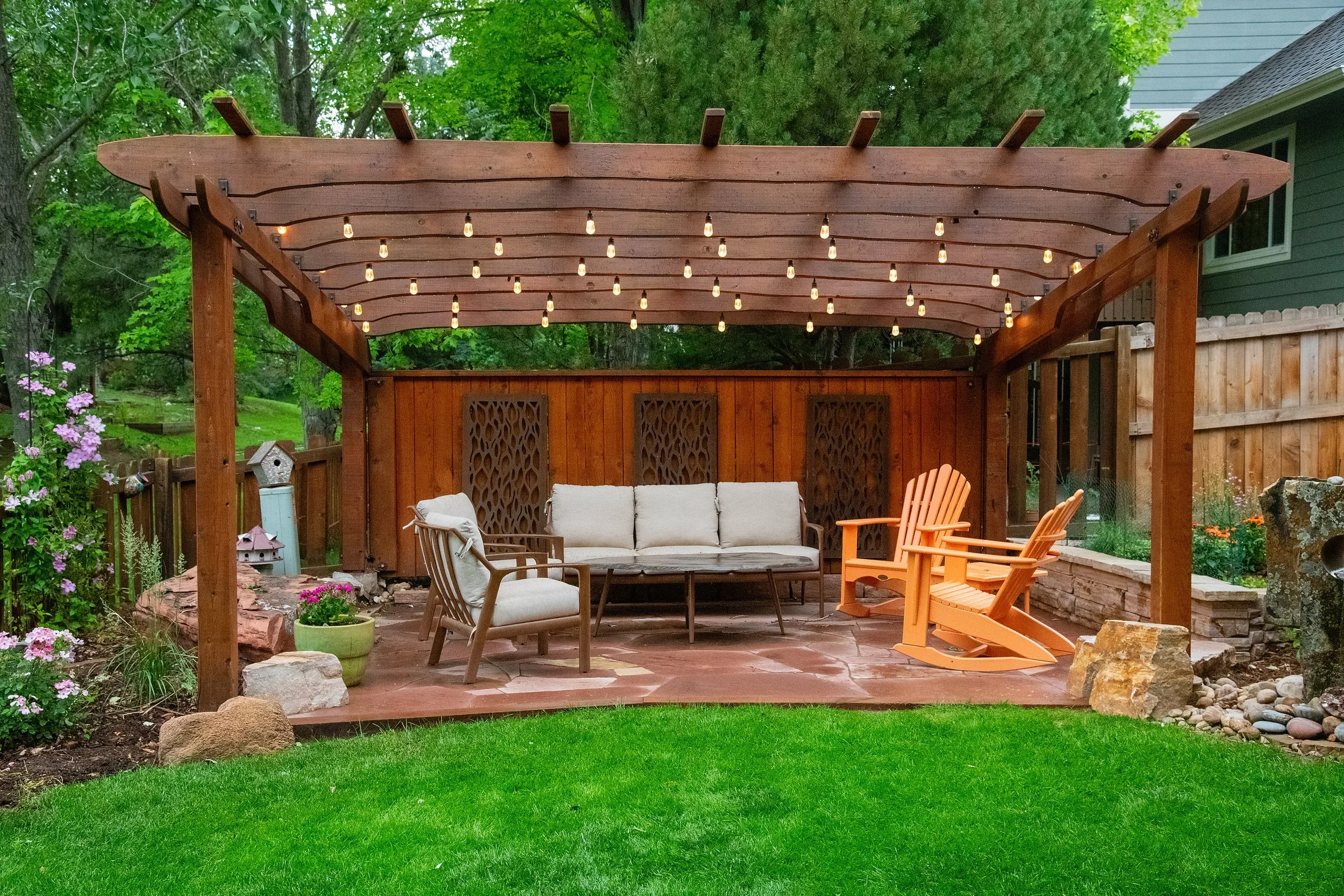 A backyard patio with a wooden pergola decorated with string lights, outdoor furniture including a cushioned sofa, chairs, and orange wooden rocking chairs, surrounded by a green lawn, flowers, rocks, and a wooden fence.
