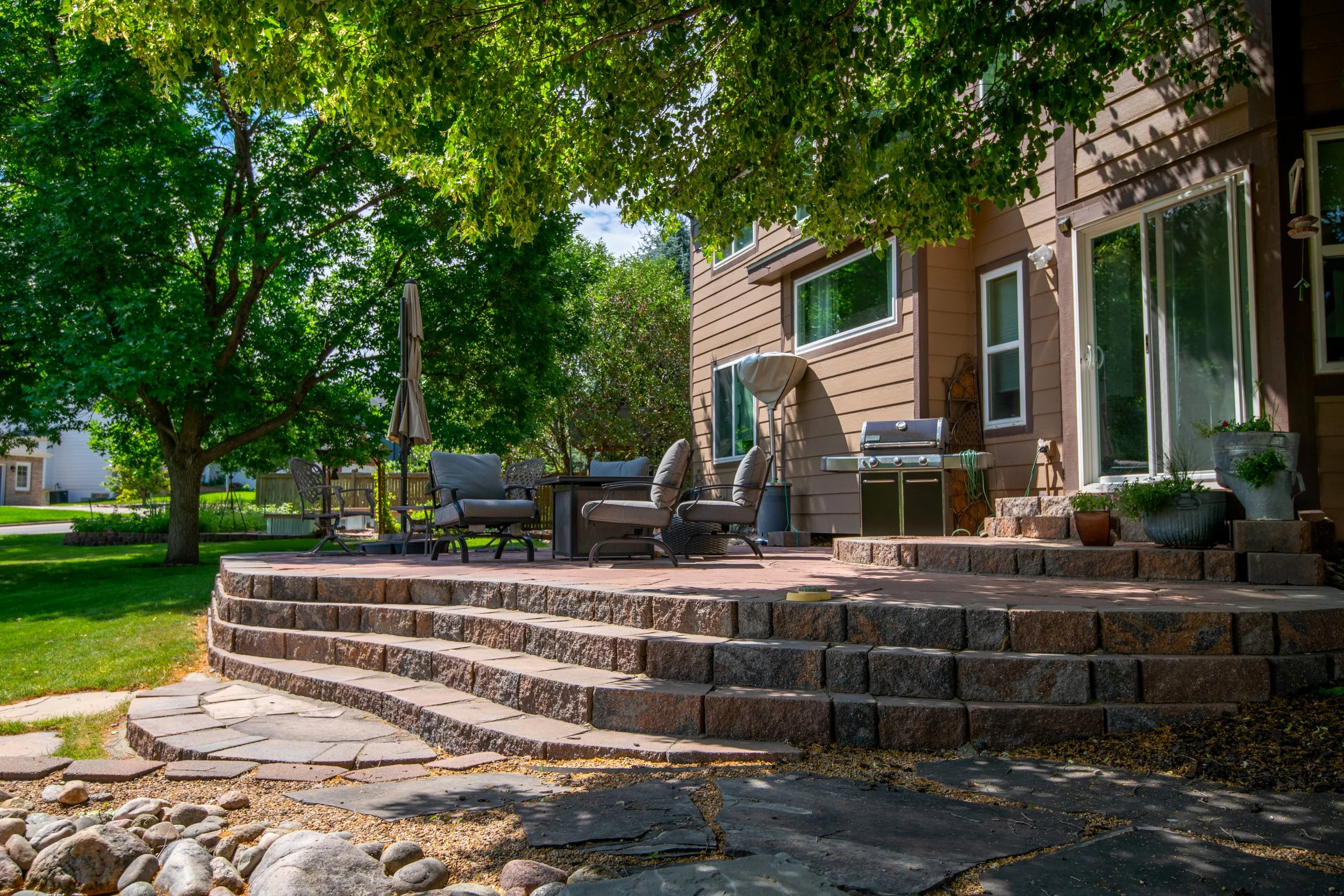Backyard with a stone patio, outdoor furniture, potted plants, grill, large tree, and a brown house with sliding glass door.