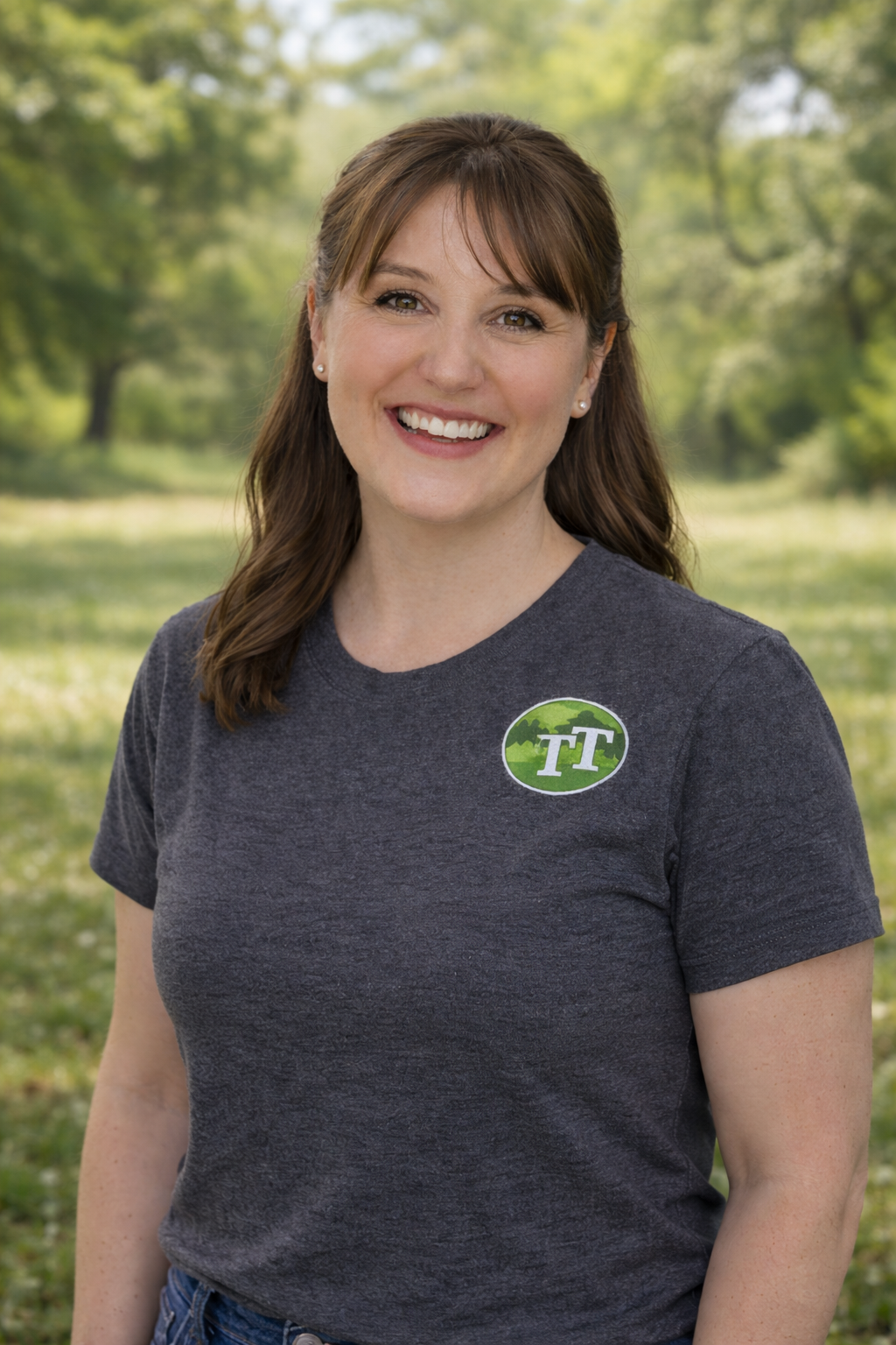 A woman smiling outdoors wearing a gray T-shirt with a logo on the chest, in a park with green trees and grass in the background.