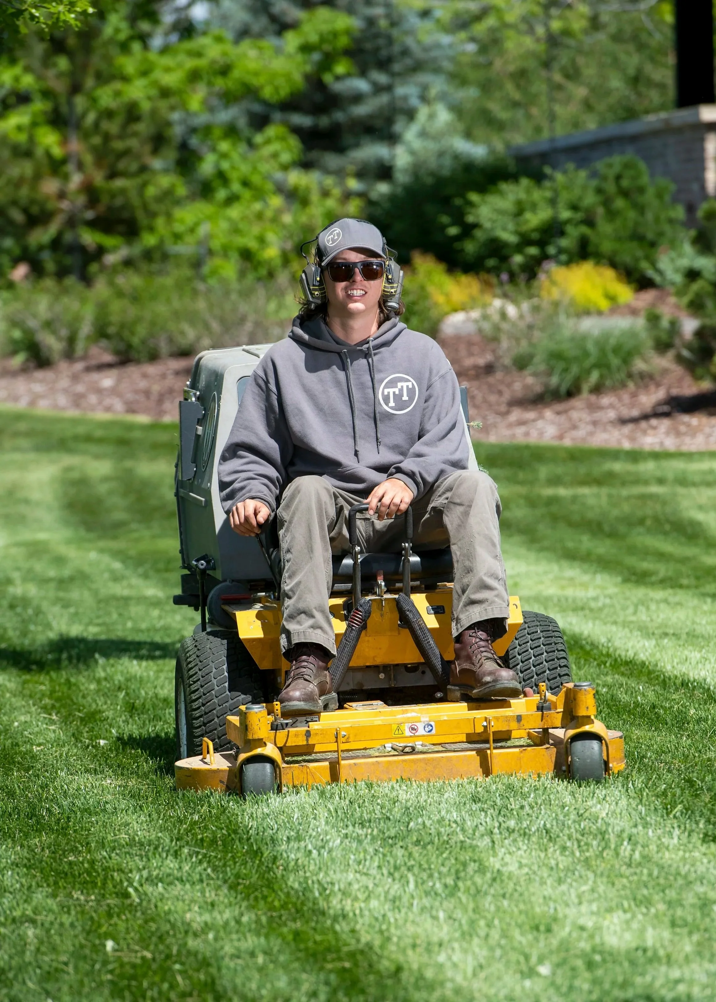 Person riding a yellow ride-on lawn mower on a well-manicured lawn, wearing sunglasses, a gray hoodie, and headphones.