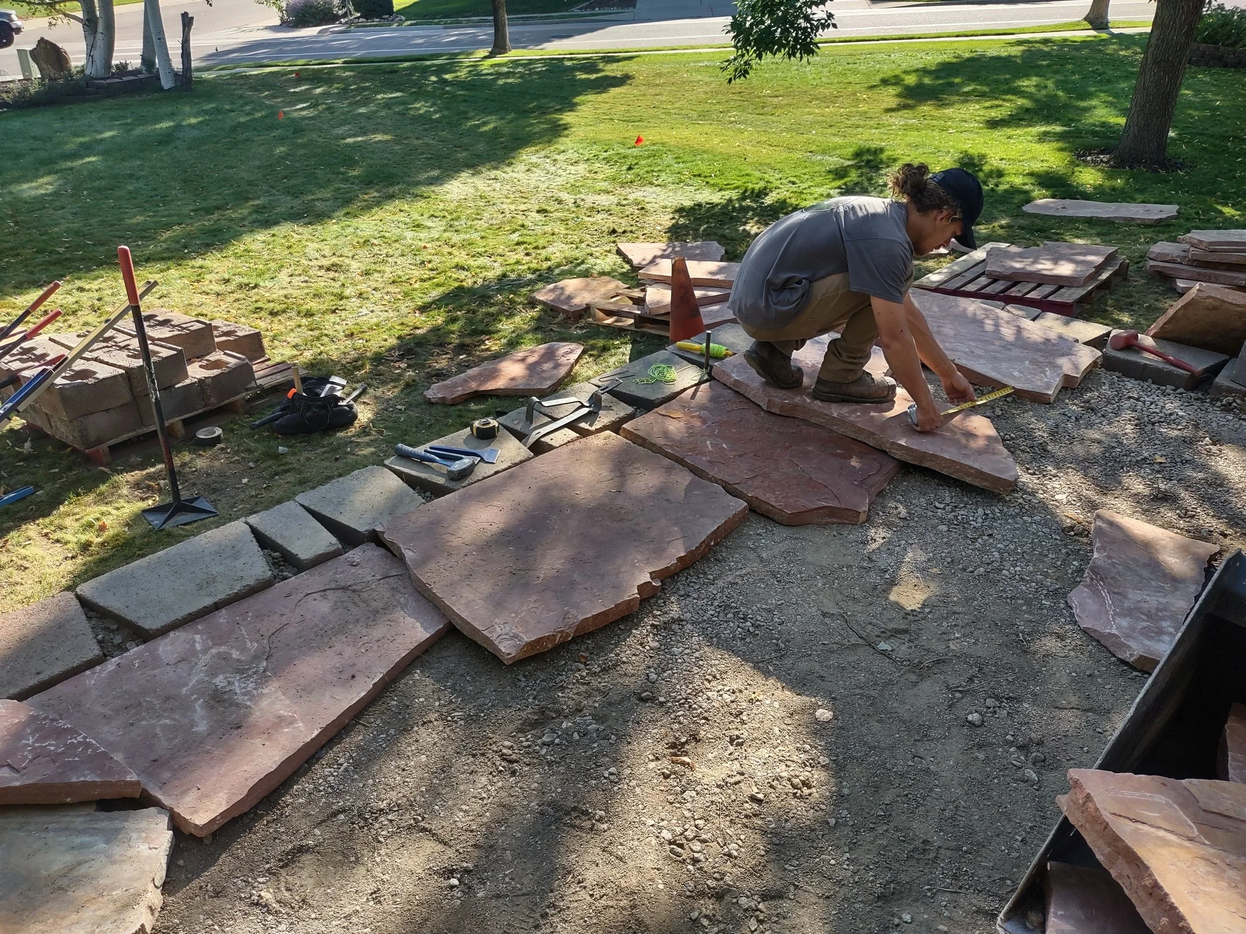 A worker installing flagstone pavers outdoors, measuring and positioning stones to create a pathway, with tools and materials nearby, in a grassy area with trees.