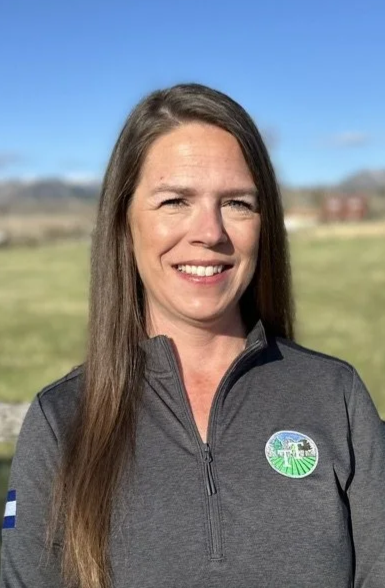 A woman with long brown hair smiling outdoors in front of a grassy field and blue sky, wearing a dark gray zip-up jacket with a circular logo on the chest.