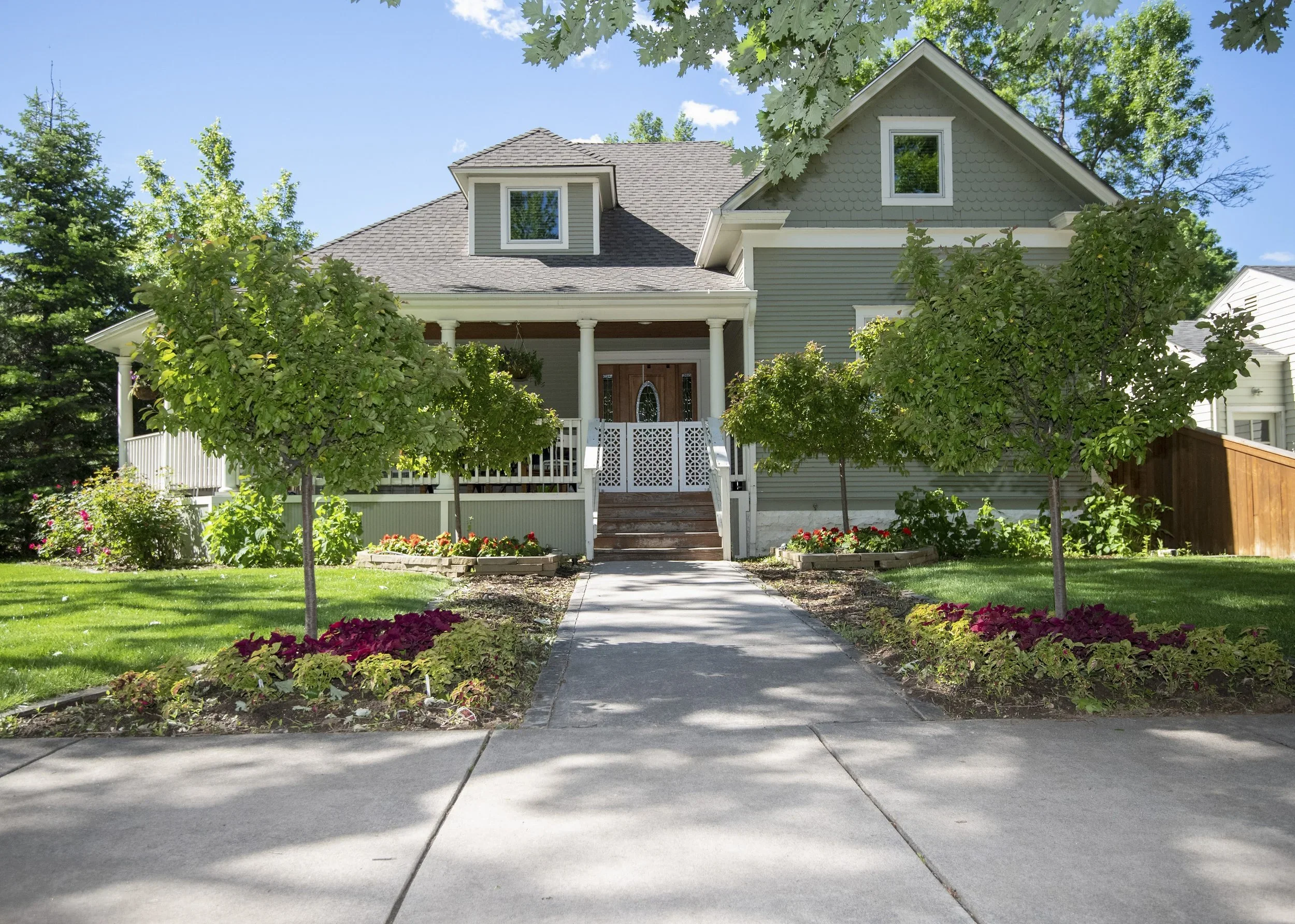 Front view of a house with a porch, surrounded by a well-maintained lawn and flower beds, with trees lining the sidewalk and a driveway in the foreground.