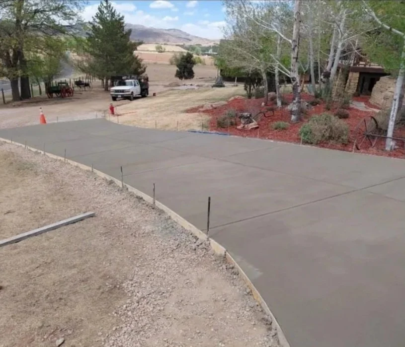 Freshly poured concrete driveway with stakes along the edges, in a rural area with trees, a pickup truck, and a building nearby.