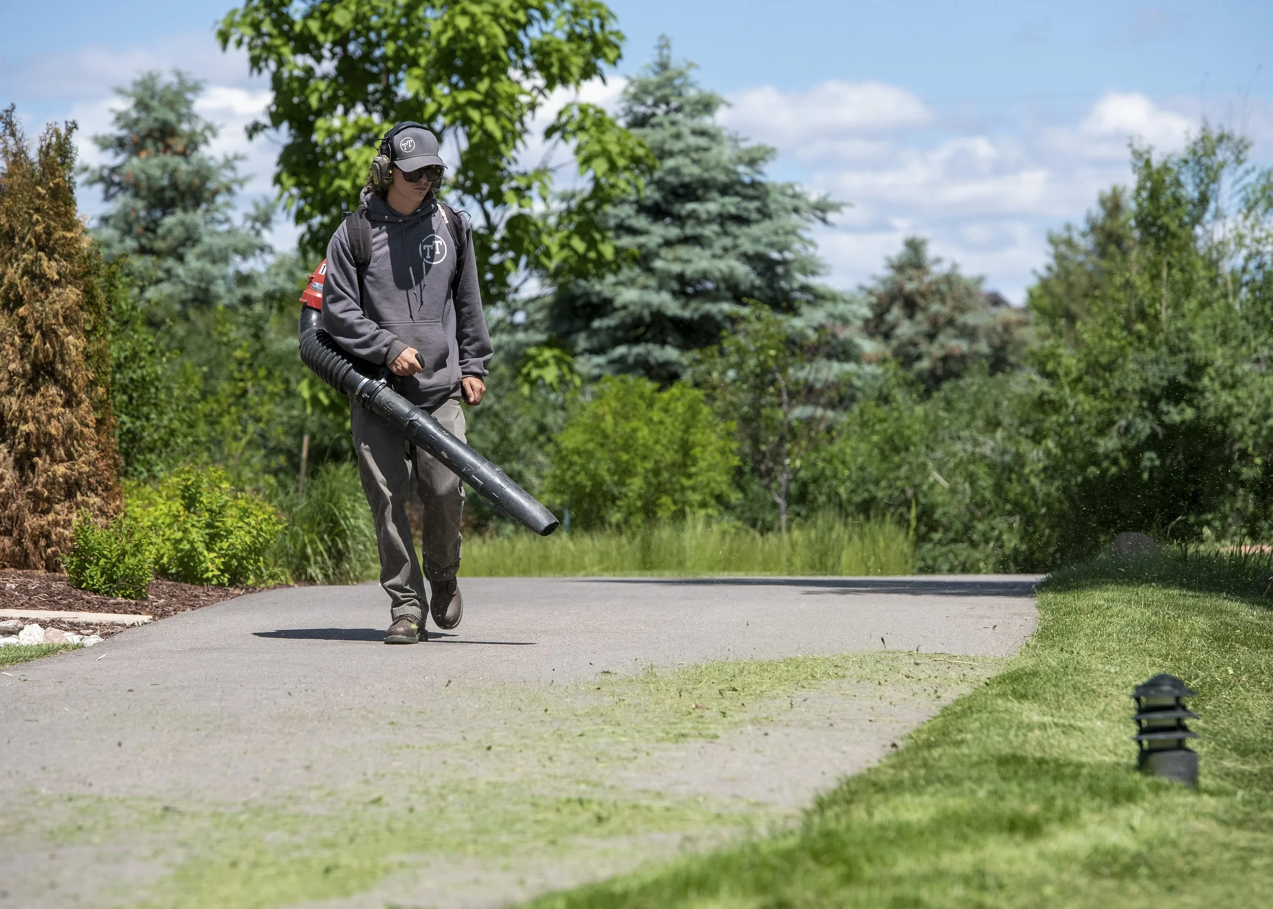 Person in gray jacket with headphones and sunglasses using leaf blower on a paved path outdoors with trees and shrubs in background.