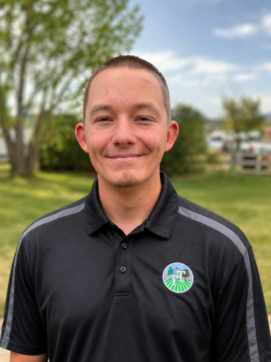 A smiling young man with short hair and a goatee wearing a black shirt with a logo on the chest, outdoors in a grassy area with trees and cars in the background.