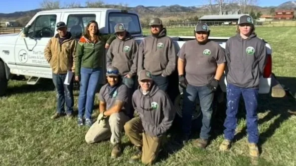 Group of eight people, including children, posing outdoors in front of a white vehicle on a grassy area with a rural landscape in the background, some wearing matching gray shirts and hats.
