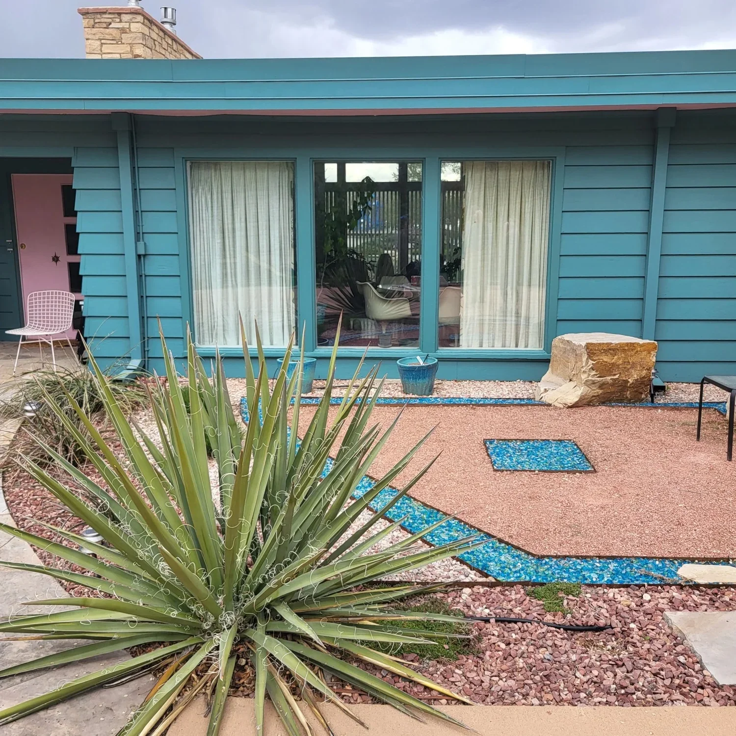 A blue house with a gravel and stone yard, large glass sliding doors, a houseplant inside, a large rock, blue planters, and outdoor furniture including a pink chair and a black chair.